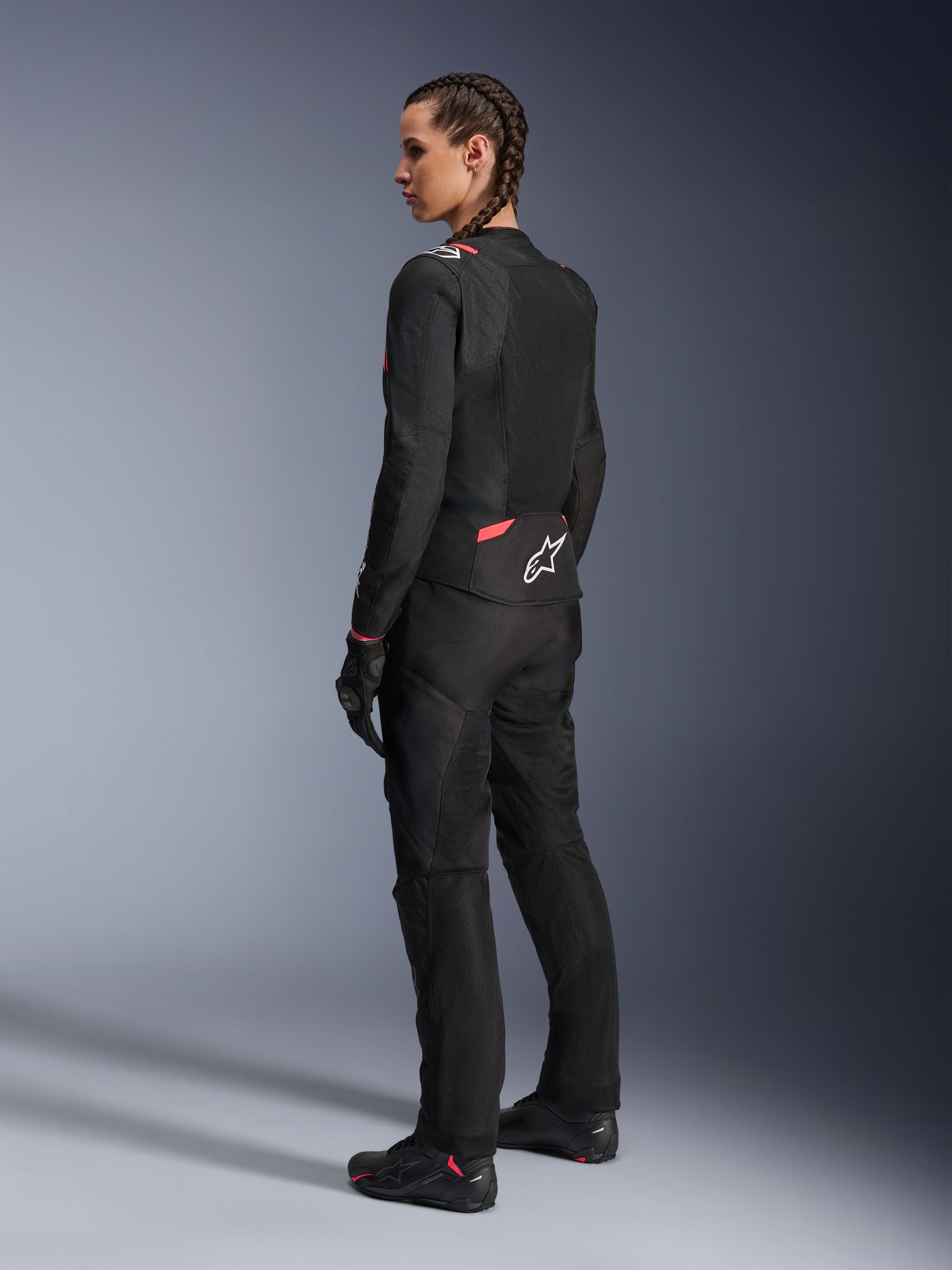 Veste Alpinestars Stella T-SPS Air v2 pour femme, noir et rose diva, veste de moto en textile vue de trois-quarts arrière, dotée de grands panneaux en mesh pour la ventilation et d'un logo étoile blanc au dos, portée avec un pantalon, des gants et des bottes de moto noirs assortis.