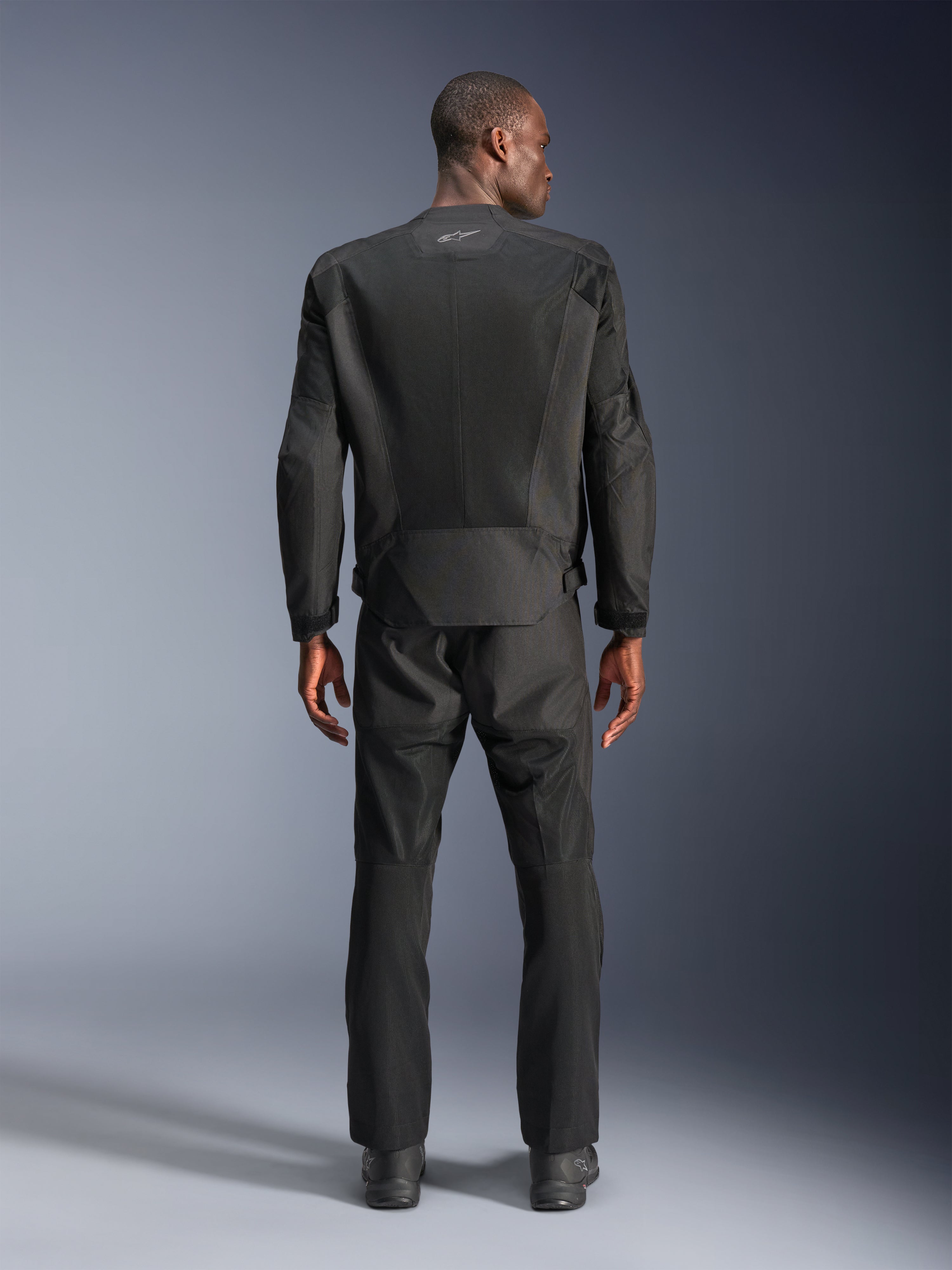Vue arrière d'un homme portant la veste Alpinestars C-1 Air en noir, veste de moto urbaine avec empiècements en maille technique, pantalon textile et bottes noirs assortis, esthétique épurée avec logo réfléchissant sur le haut du dos.