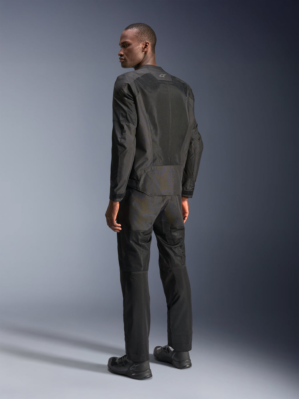 Veste Alpinestars C-1 Air, veste de moto urbaine, noire, vue arrière montrant les panneaux en maille technique pour la circulation de l'air et le logo réfléchissant, portée avec un pantalon et des chaussures de moto noirs assortis, esthétique épurée et raffinée pour les trajets en ville