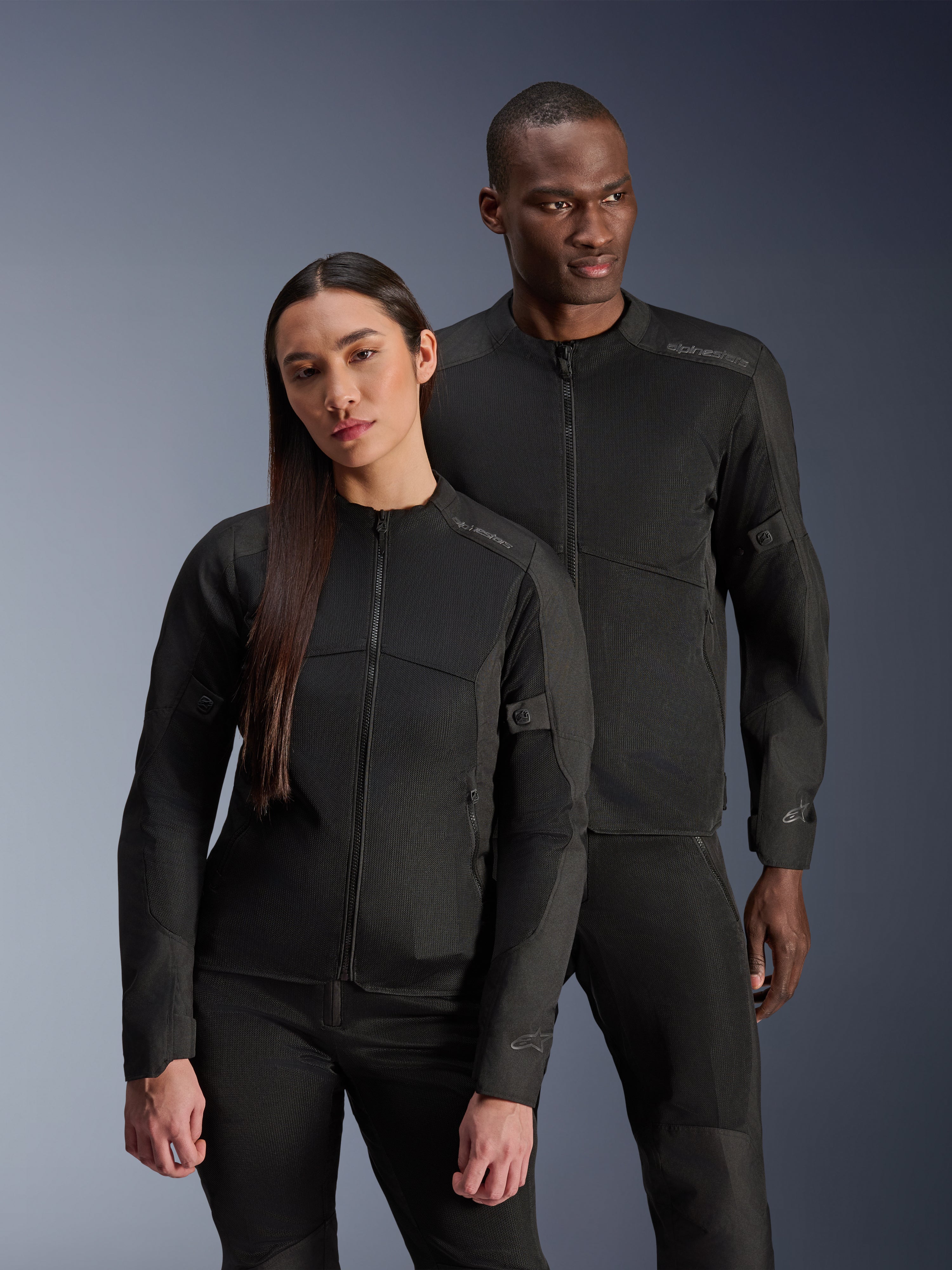 Un homme et une femme portant les vestes Alpinestars C-1 Air et Stella C-1 Air, vestes de moto urbaines assorties en textile noir avec panneaux en mesh pour la ventilation, associées à des pantalons de moto noirs pour un look contemporain et épuré.