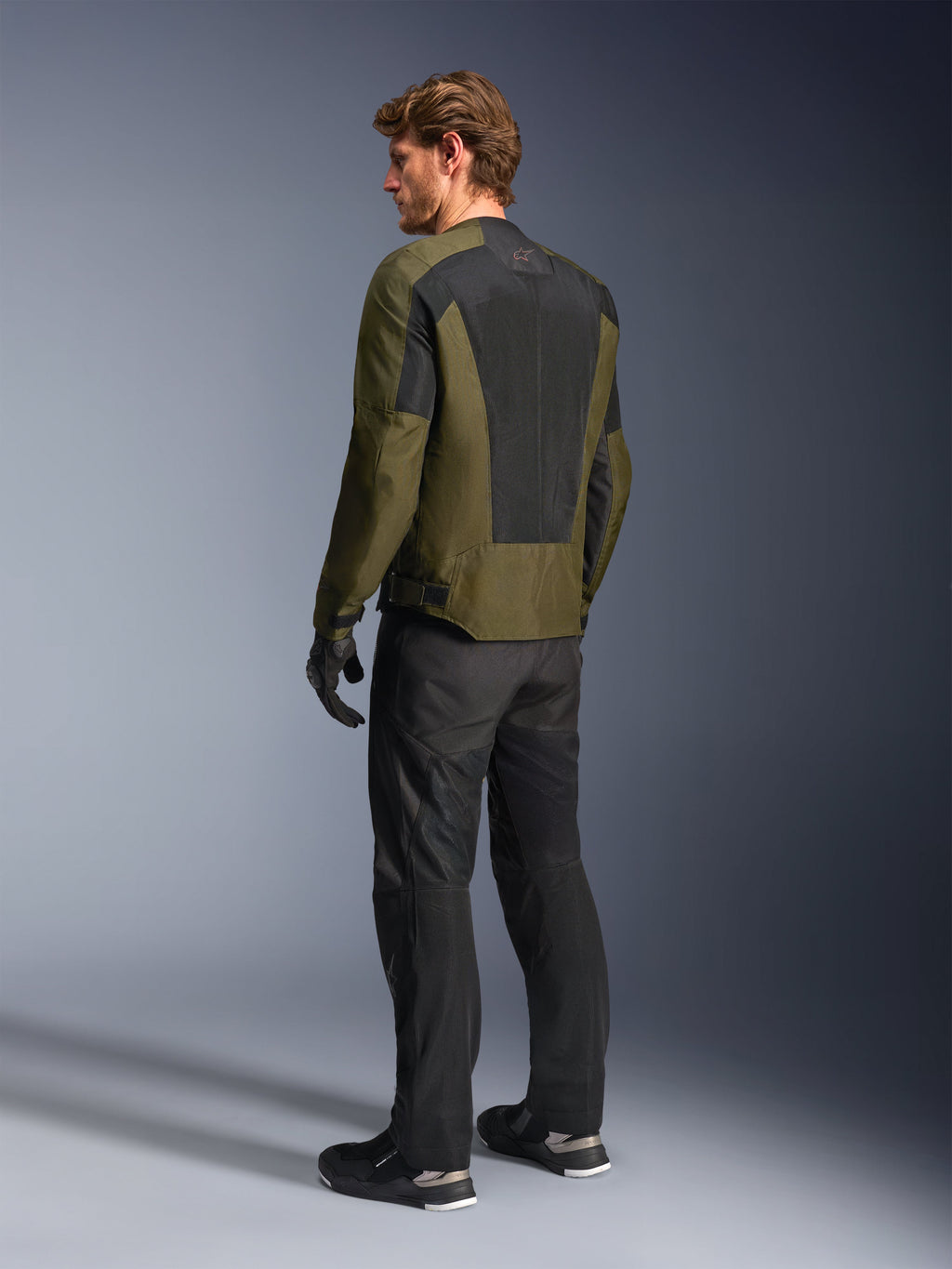 Modèle masculin portant la veste Alpinestars C-1 Air en Khaki, veste de moto urbaine, vue arrière mettant en évidence de larges panneaux en maille noire dans le dos, associée à un pantalon de moto technique et des gants noirs, équipement de moto contemporain pour temps chaud