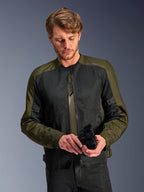 Homme portant la veste Alpinestars C-1 Air en Khaki, veste de moto urbaine avec textile vert olive et panneaux en maille noire, regardant vers le bas tout en tenant des gants de moto noirs, style épuré et contemporain pour la conduite sur route.