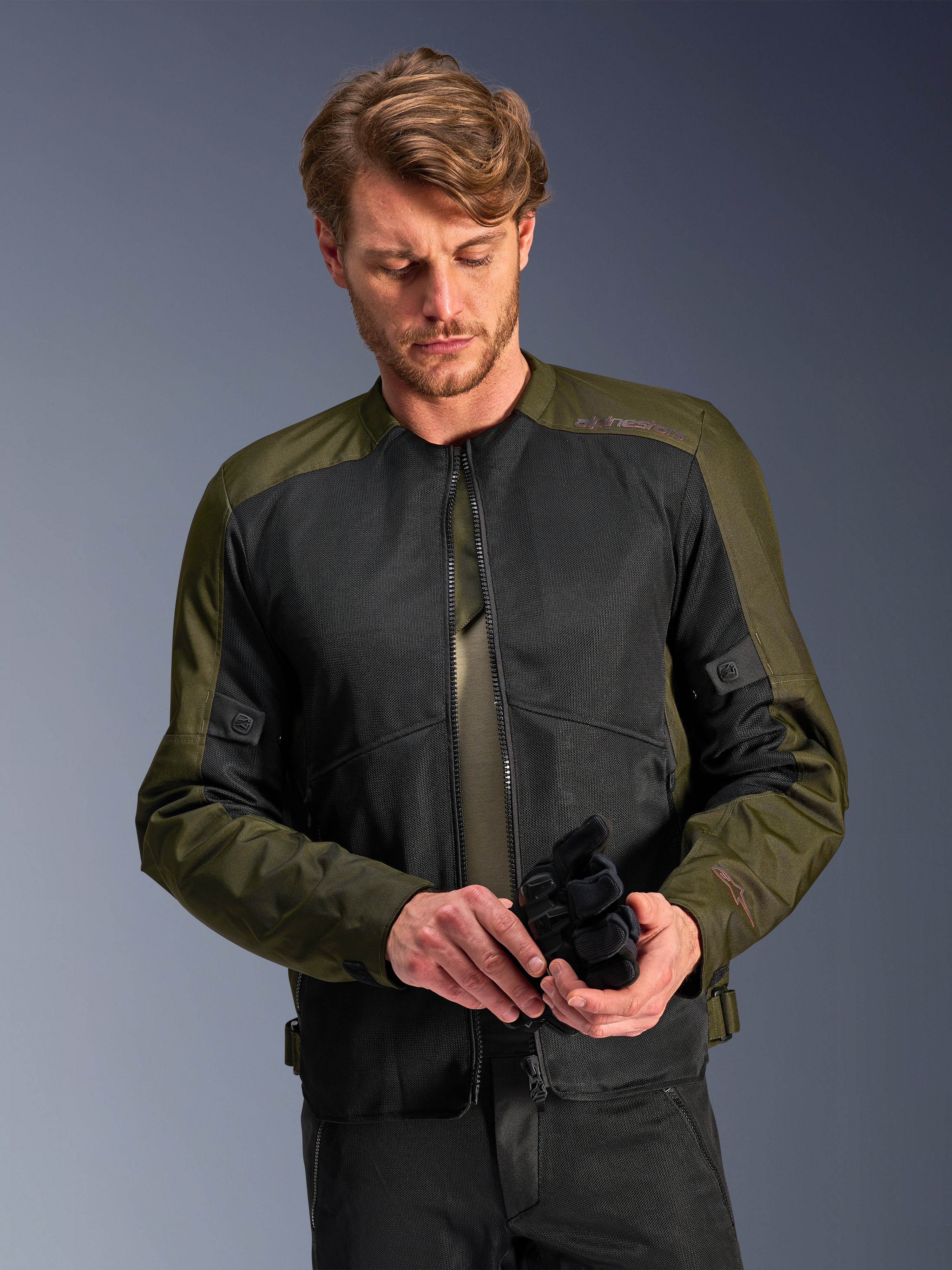 Homme portant la veste Alpinestars C-1 Air en Khaki, veste de moto urbaine avec textile vert olive et panneaux en maille noire, regardant vers le bas tout en tenant des gants de moto noirs, style épuré et contemporain pour la conduite sur route.