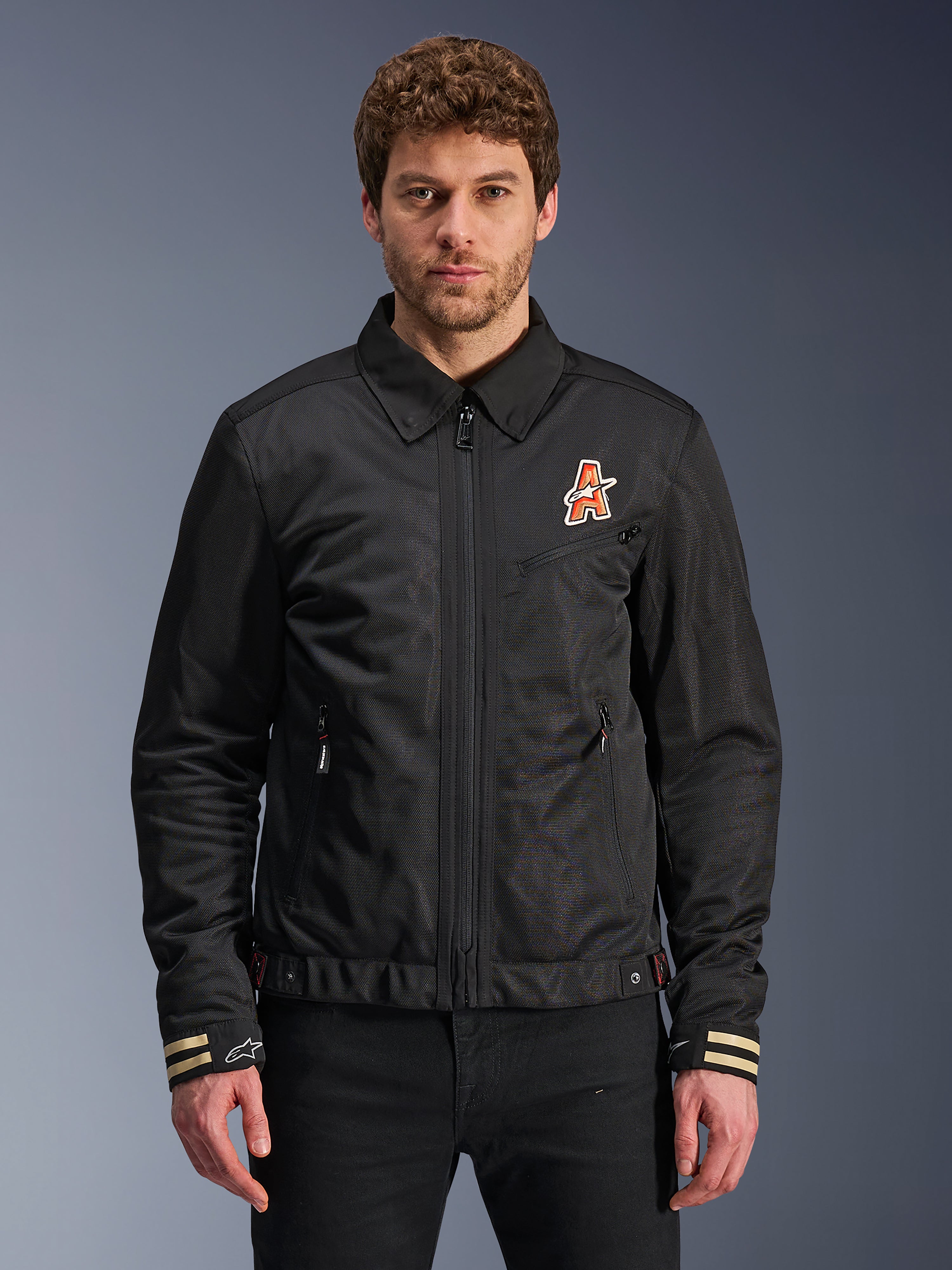 Alpinestars Ignite Air Jacket, Black motorcycle jacket, traditional shirt collar, orange and white 'A' logo on chest, beige striped cuffs with white star logo, urban contemporary riding gear