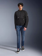 Modèle masculin portant la veste Alpinestars Provoke en noir, veste de moto urbaine en textile dotée de fermetures éclair techniques et d'inserts en softshell ripstop, associée à un jean bleu et des baskets noires, style de motard urbain contemporain.