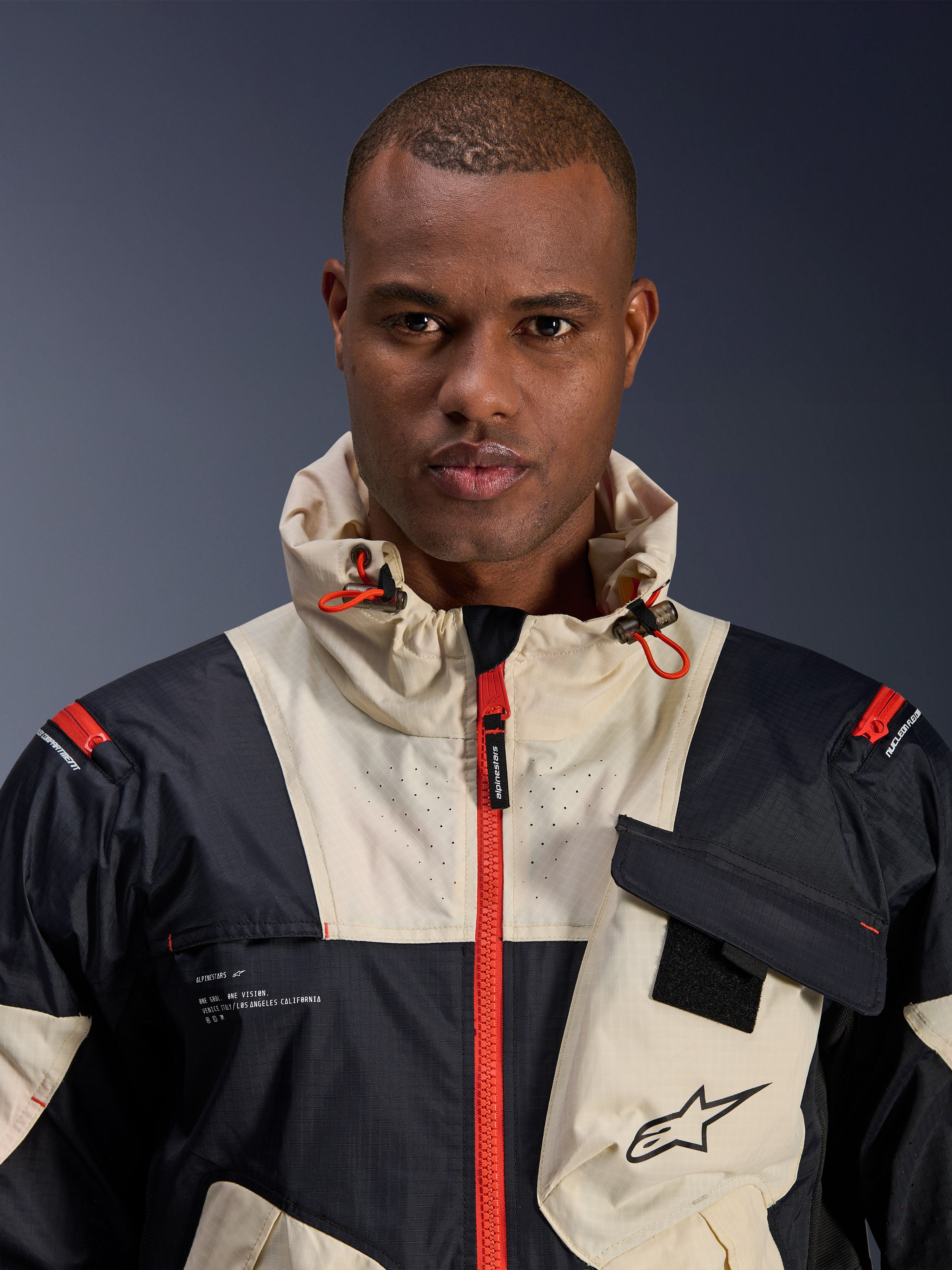 Male model wearing Alpinestars Mogress Air Jacket, Urban Motorcycle Jacket, Black Almond and Hot Orange, close-up view showing the color-blocked design, ripstop texture, hooded collar with orange toggles, and front zipper details.
