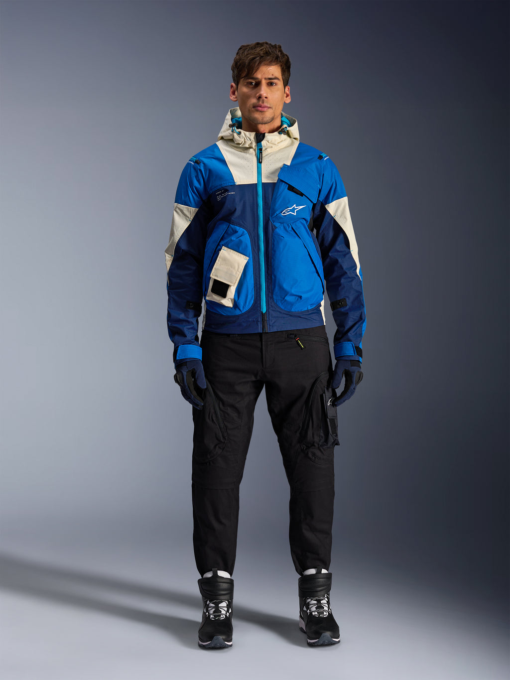 Veste Alpinestars Mogress Air, veste de moto urbaine Bleu Amande, portée avec un pantalon et des bottes noirs, présentant un design technique color-block bleu et crème avec des poches utilitaires et une capuche, vêtement de moto contemporain d'inspiration streetwear