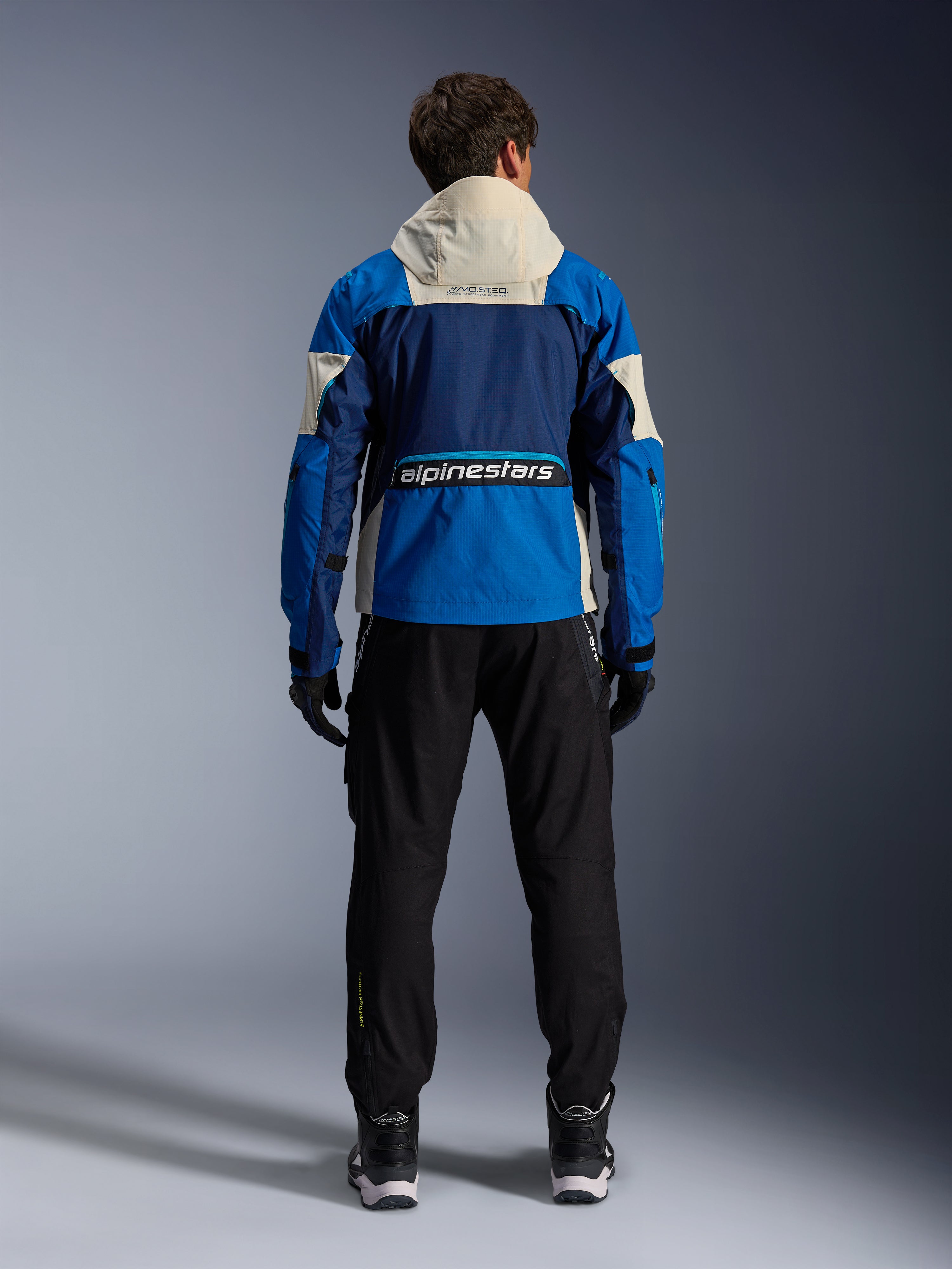 Veste Alpinestars Mogress Air en Blue Almond, vue arrière d'un motard portant la veste de moto, présentant des empiècements bleu marine et bleu vif et une capuche blanc cassé, logo Alpinestars imposant dans le bas du dos, style urbain contemporain.