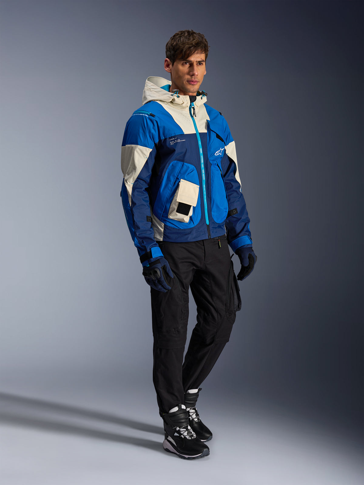 Veste Alpinestars Mogress Air, veste de moto urbaine, coloris Bleu Amande avec empiècements crème et bleu marine, dotée d'une capuche et de poches utilitaires, portée avec un pantalon de moto noir, des gants et des bottes, équipement de protection contemporain inspiré du streetwear