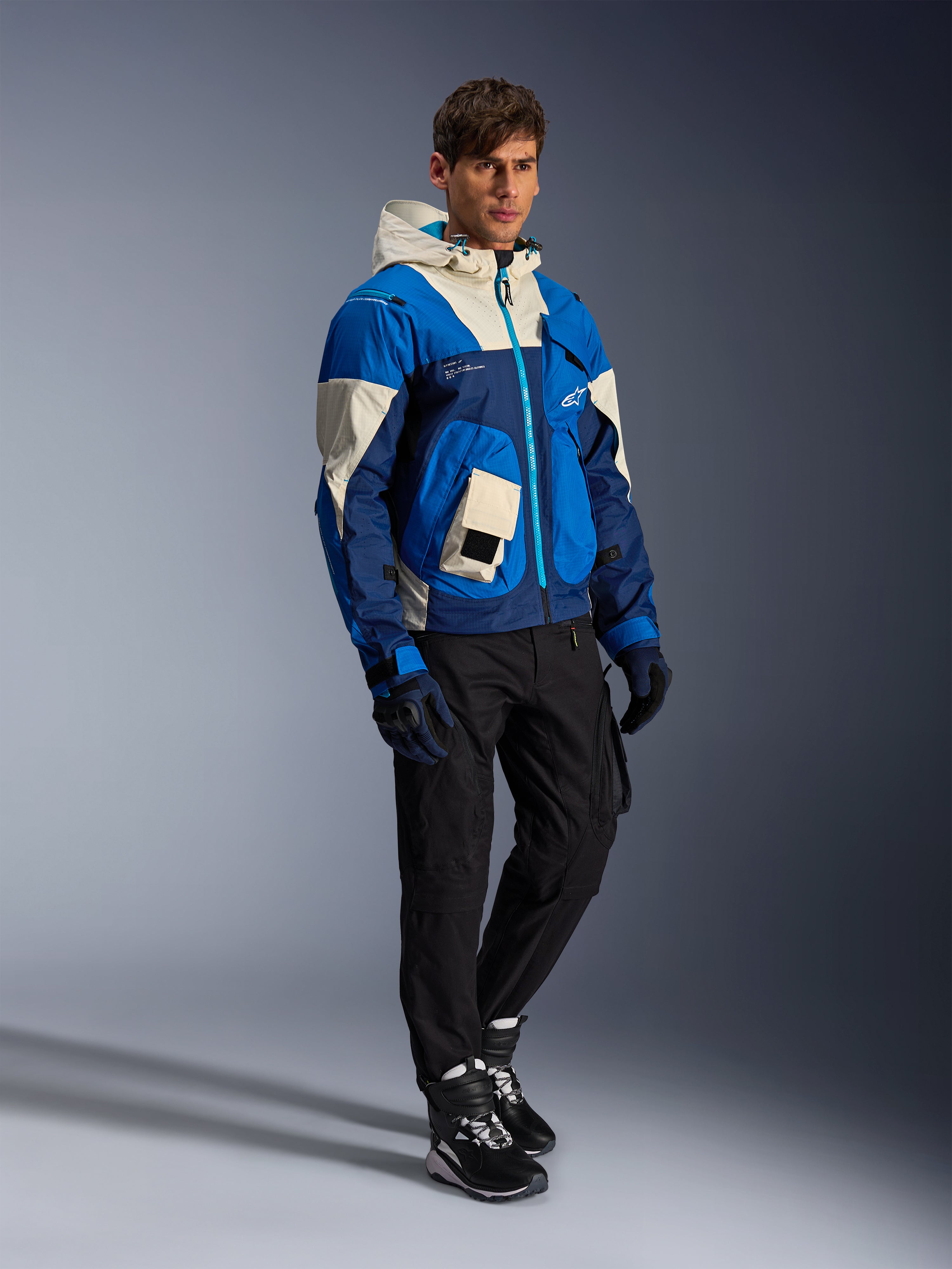 Veste Alpinestars Mogress Air, veste de moto urbaine, coloris Bleu Amande avec empiècements crème et bleu marine, dotée d'une capuche et de poches utilitaires, portée avec un pantalon de moto noir, des gants et des bottes, équipement de protection contemporain inspiré du streetwear