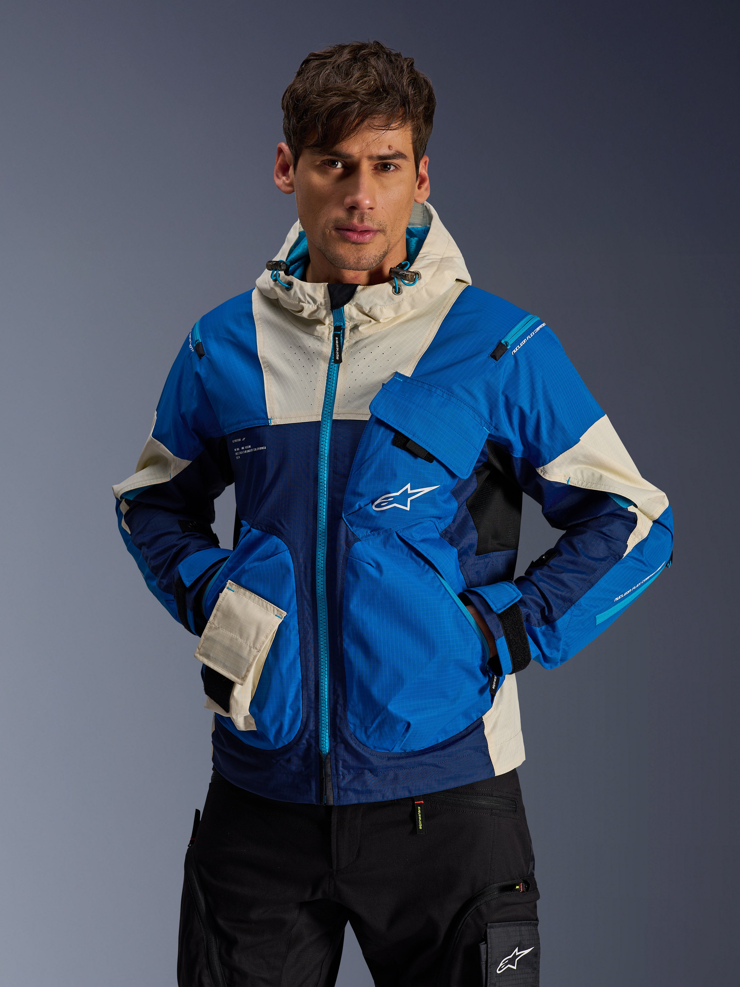 Modèle masculin portant la veste Alpinestars Mogress Air en Blue Almond, veste de moto urbaine avec blocs de couleurs bleu vif et bleu marine, accents crème sur la capuche et les poches, tissu technique ripstop avec poches utilitaires, streetwear moderne inspiré du sport automobile