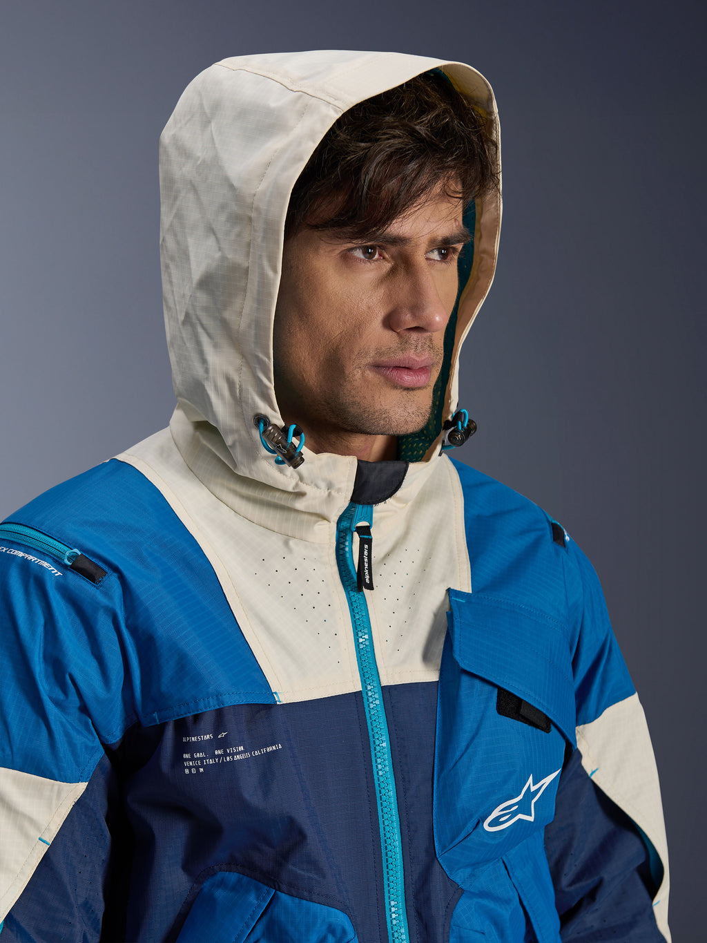 Veste Alpinestars Mogress Air en Blue Almond, veste de moto urbaine présentant un design color-block en bleu et crème, mannequin portant la capuche relevée, conception textile légère avec détails de fermetures éclair techniques.