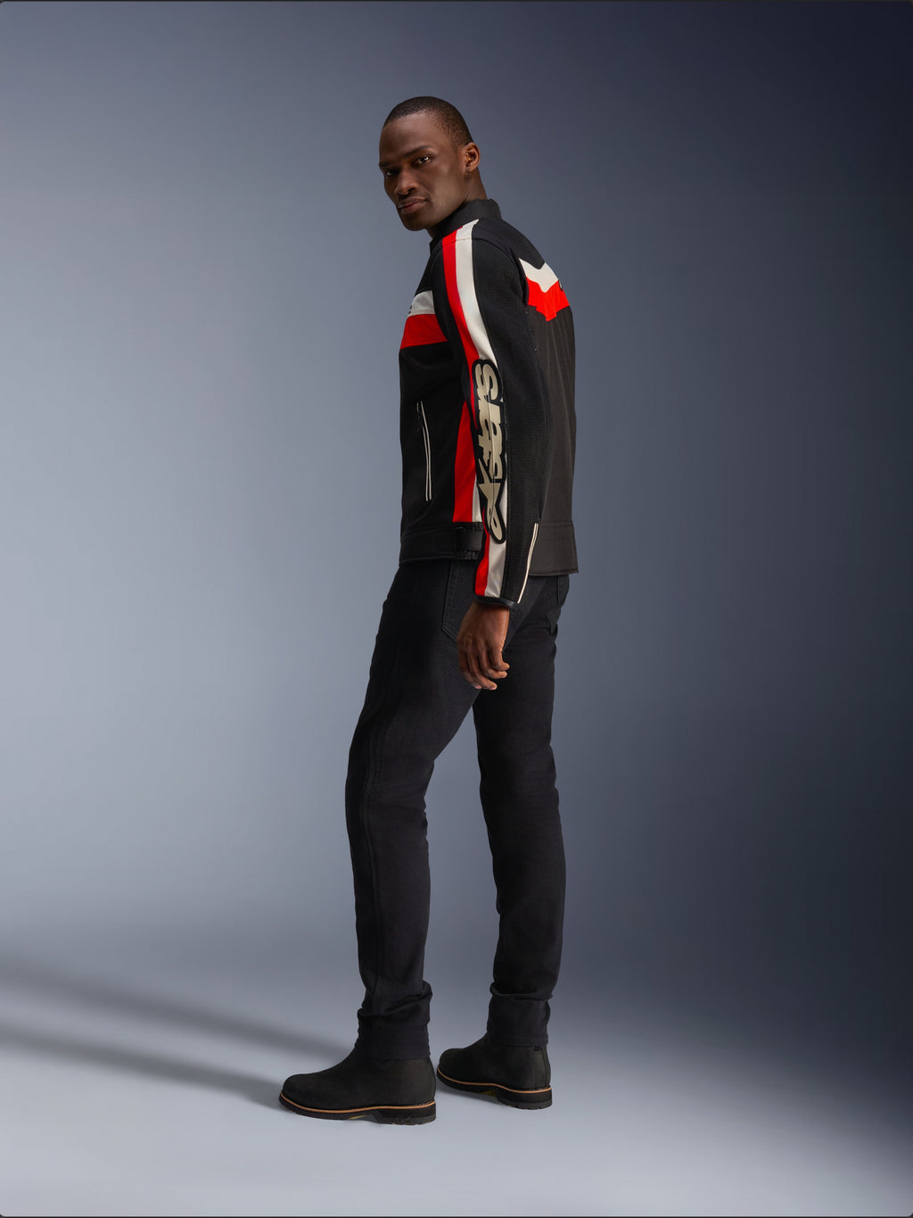 Un homme portant la veste Alpinestars T-Dyno Air en noir, rouge vif et écru, veste de moto en textile d'inspiration vintage avec des panneaux en maille, logo imposant sur la manche, associée à un pantalon et des bottes noirs pour un look motard moderne