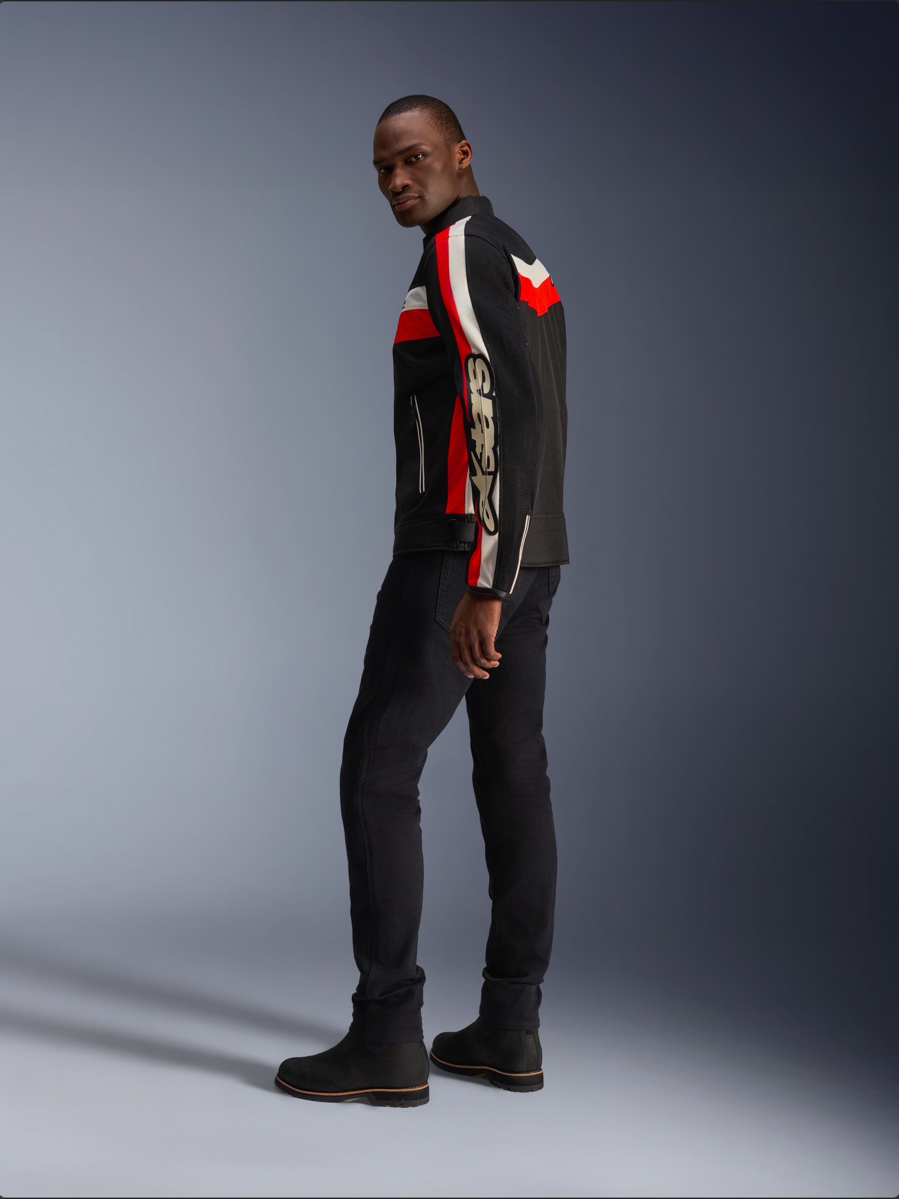 Un homme portant la veste Alpinestars T-Dyno Air en noir, rouge vif et écru, veste de moto en textile d'inspiration vintage avec des panneaux en maille, logo imposant sur la manche, associée à un pantalon et des bottes noirs pour un look motard moderne