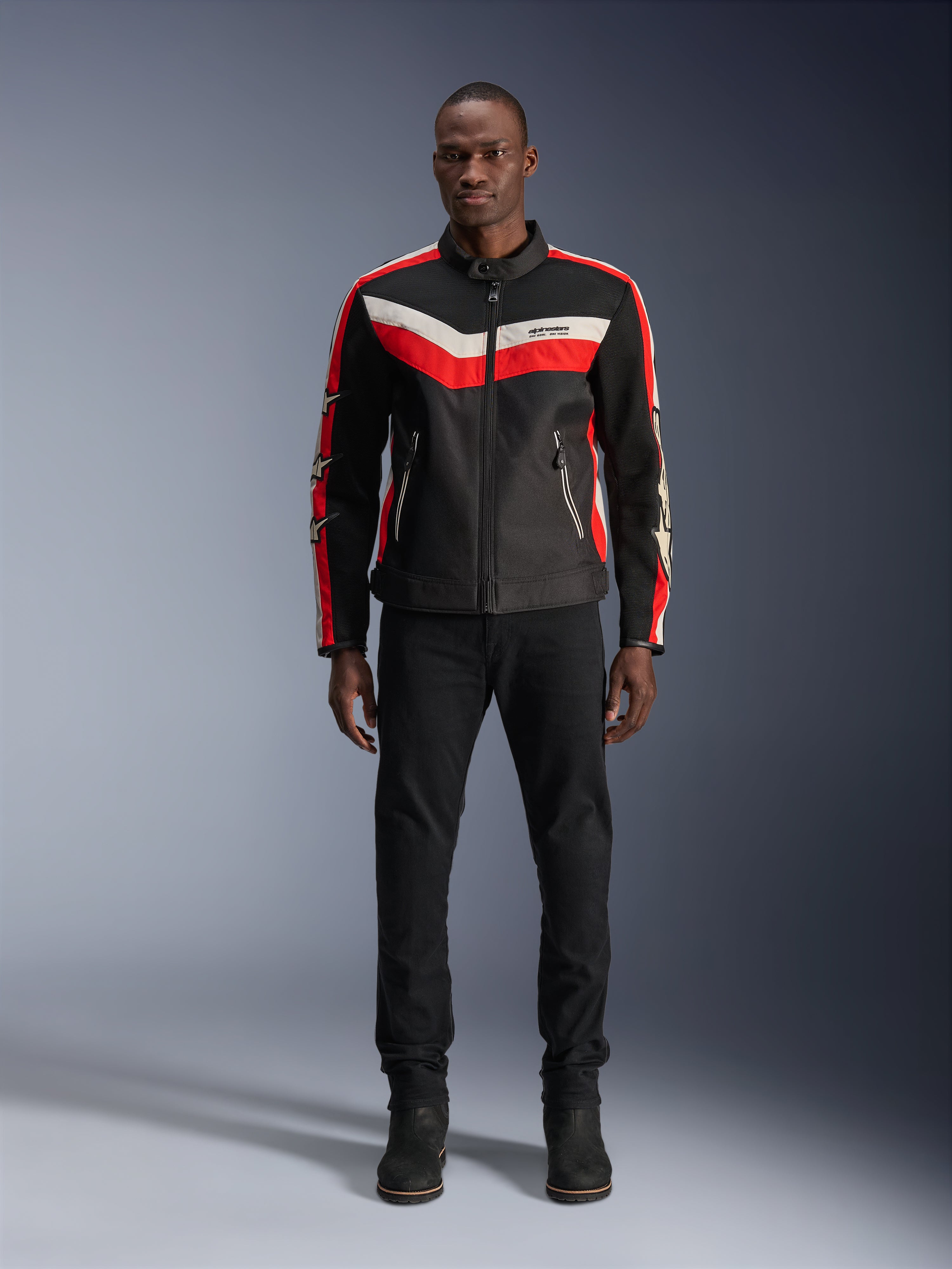 Veste Alpinestars T-Dyno Air, veste de moto en textile, noir, rouge vif et écru, portée par un mannequin avec un pantalon et des bottes noirs, dotée d'un design d'inspiration vintage avec des bandes de couleurs vives sur la poitrine, des logos étoiles sur les manches et une construction en maille respirante pour la conduite urbaine.