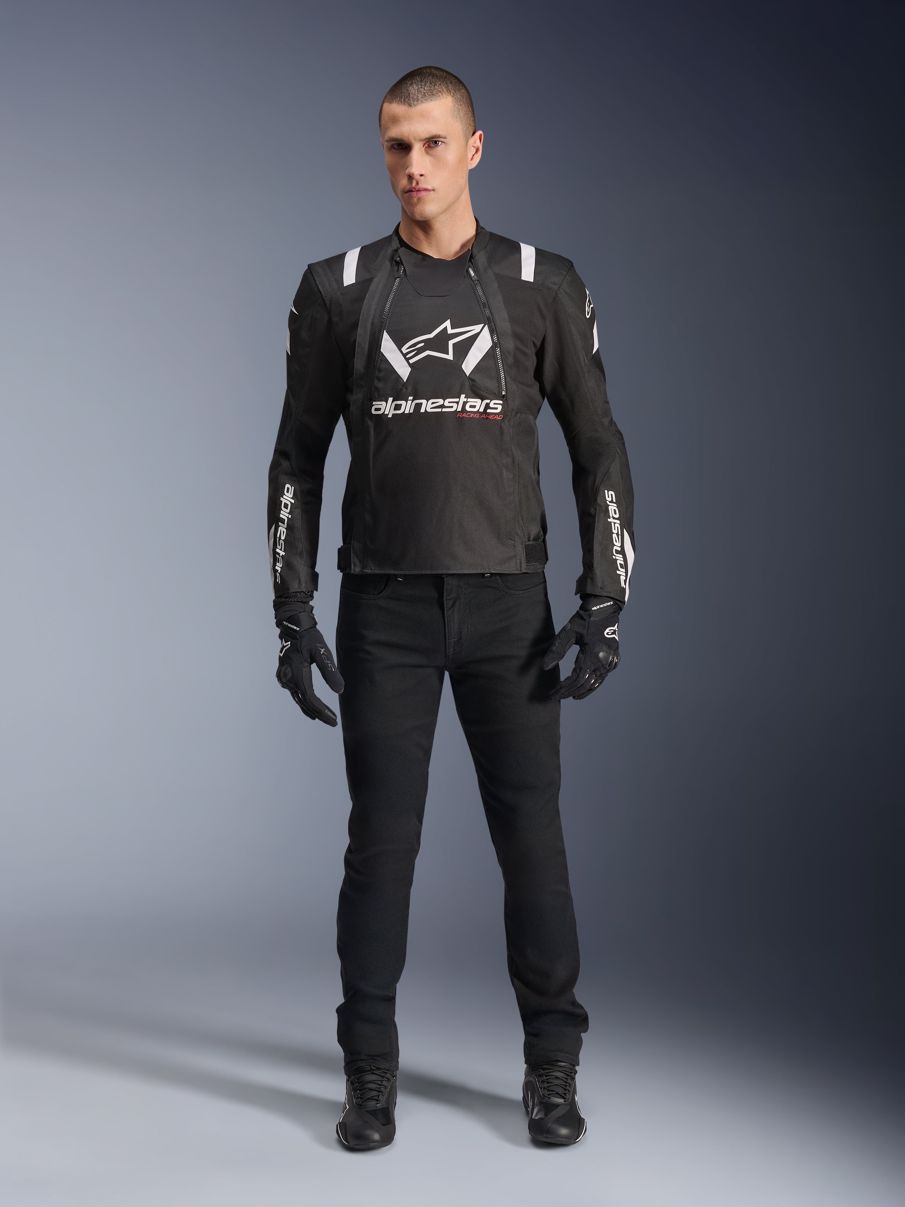 Veste Alpinestars T-Stunt Air, veste de moto noire et blanche, portée avec un pantalon de moto, des gants et des bottes noirs, logo Alpinestars blanc sur la poitrine et les manches, conception textile à coupe sportive avec panneaux en mesh pour la ventilation