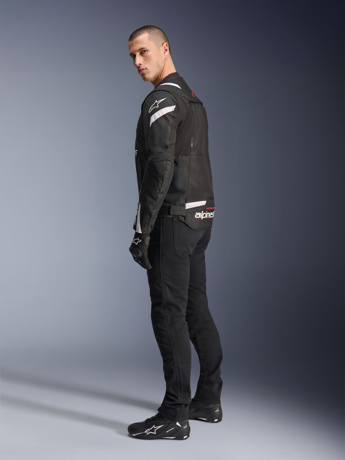 Veste Alpinestars T-Stunt Air, noir et blanc, veste de moto modulaire avec panneaux en maille technique, vue de profil arrière sur un mannequin, portée avec un pantalon de moto et des chaussures noirs, avec des accents blancs et des logos sur les épaules et le dos.