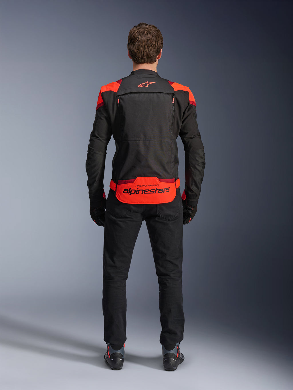 Veste Alpinestars T-Stunt Air, noire avec accents Rouge Fluo et Rouge Foncé, vue de dos sur modèle, accompagnée d'un pantalon de moto et de bottes noirs, arborant un marquage Alpinestars imposant sur le bas du dos, conception modulaire avec mesh pour la ventilation.