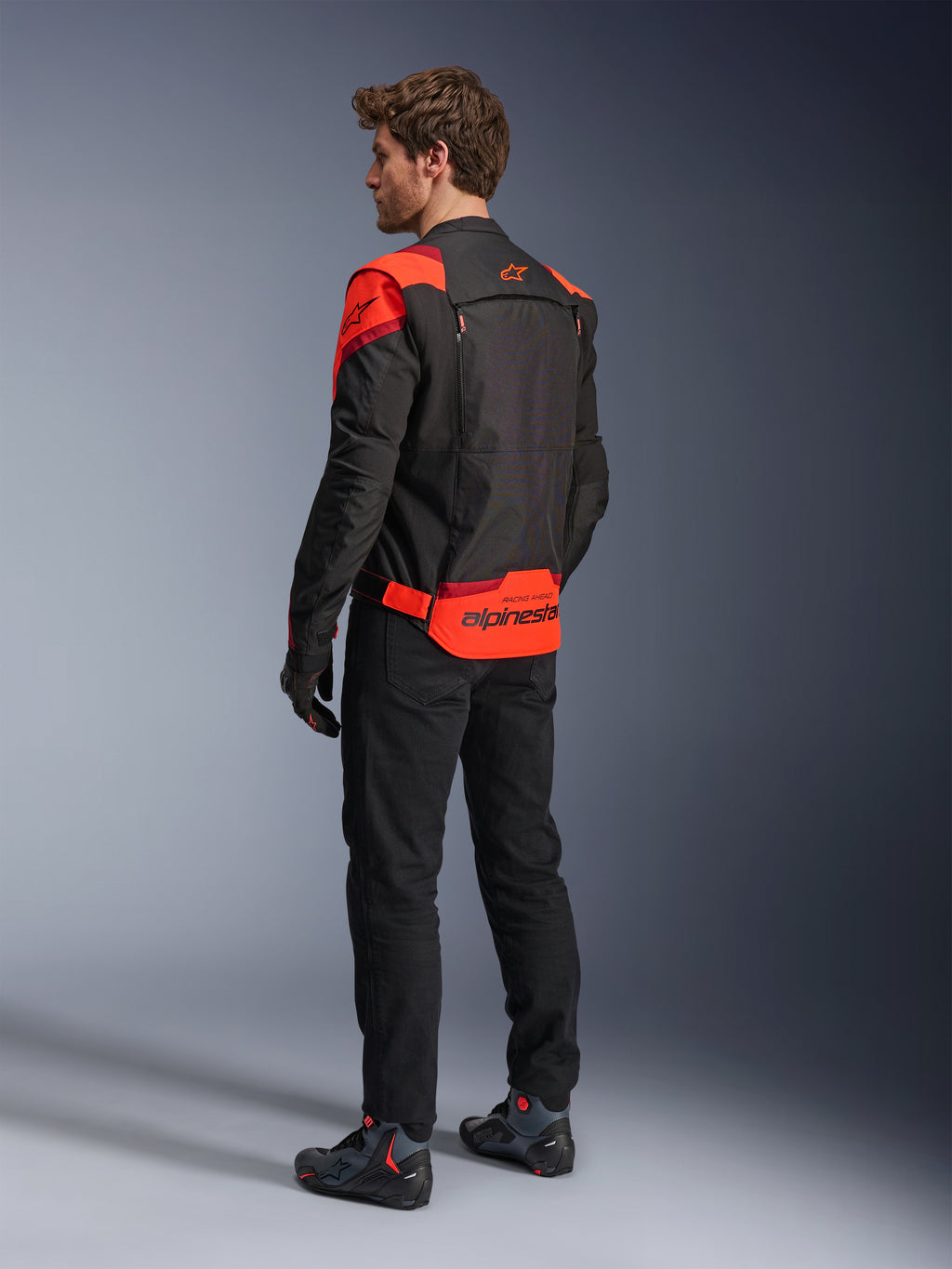 Homme portant la veste Alpinestars T-Stunt Air vue de dos, Noir Rouge Fluo et Rouge Foncé, veste de moto en textile avec panneaux en mesh et coupe sportive, avec le logo Alpinestars imposant dans le bas du dos, associée à un pantalon noir et des bottes de moto