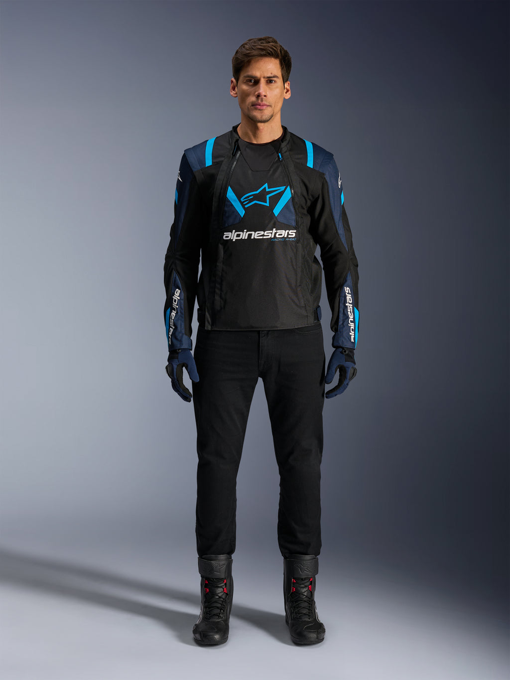 Alpinestars T-Stunt Air Jacket, veste de moto, noir avec accents bleu foncé et bleu vif, grand logo blanc sur la poitrine, construction en mesh à coupe sportive pour la circulation de l'air, portée avec un pantalon de moto noir et des bottes pour une esthétique de route épurée