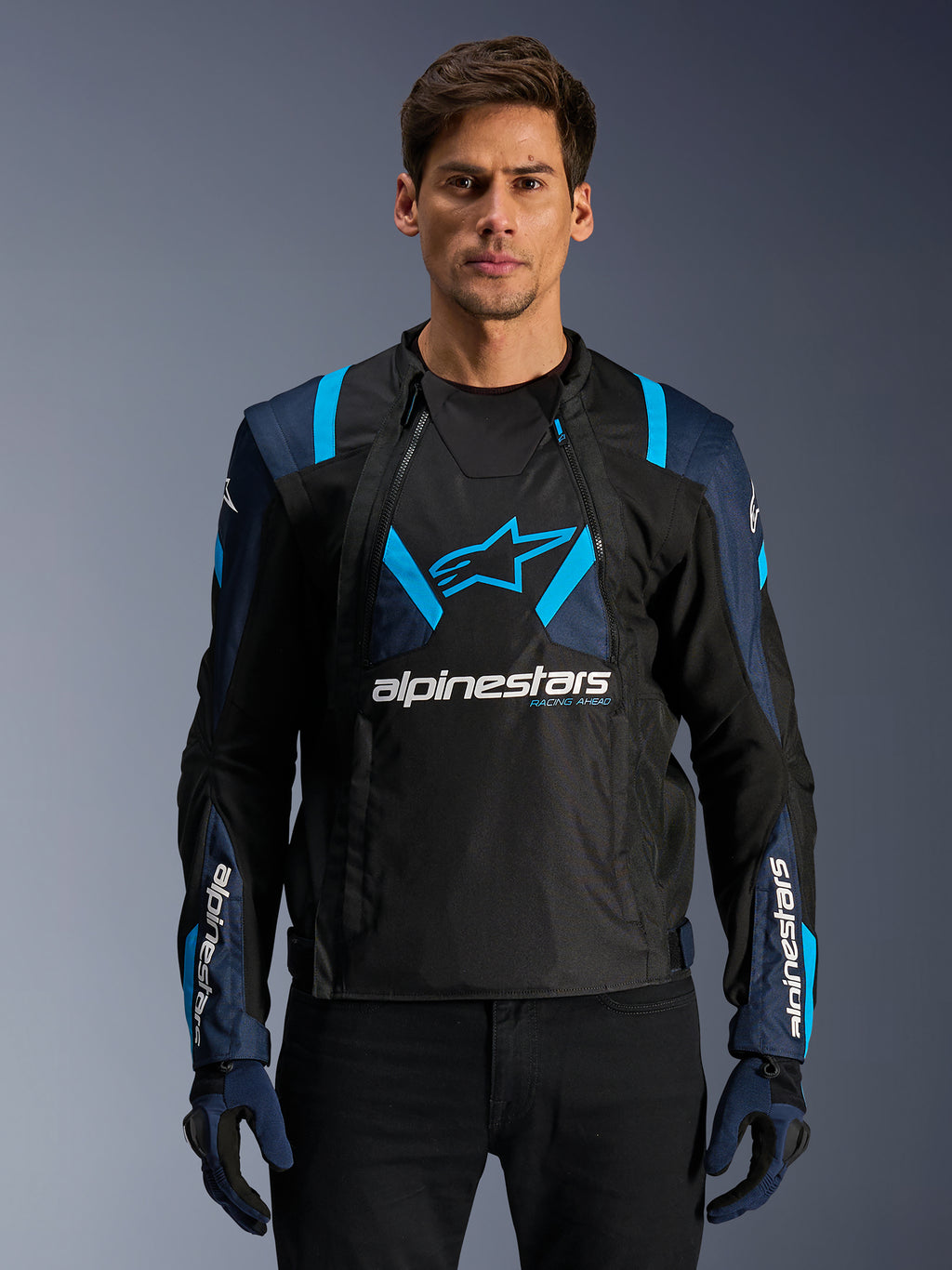Veste Alpinestars T-Stunt Air, veste de moto en mesh, noire et bleue, portée par un mannequin avec un pantalon noir et des gants, dotée d'un design modulaire 2-en-1 avec des panneaux en mesh technique respirant, logo étoile sur la poitrine et marquage Alpinestars sur les manches.