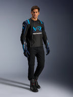Modèle masculin portant la veste Alpinestars T-Stunt Air, noir, bleu foncé et bleu vif, dotée de larges panneaux en mesh pour la circulation de l'air, associée à un pantalon noir et des gants de moto, veste de moto à coupe sportive pour la route en été.