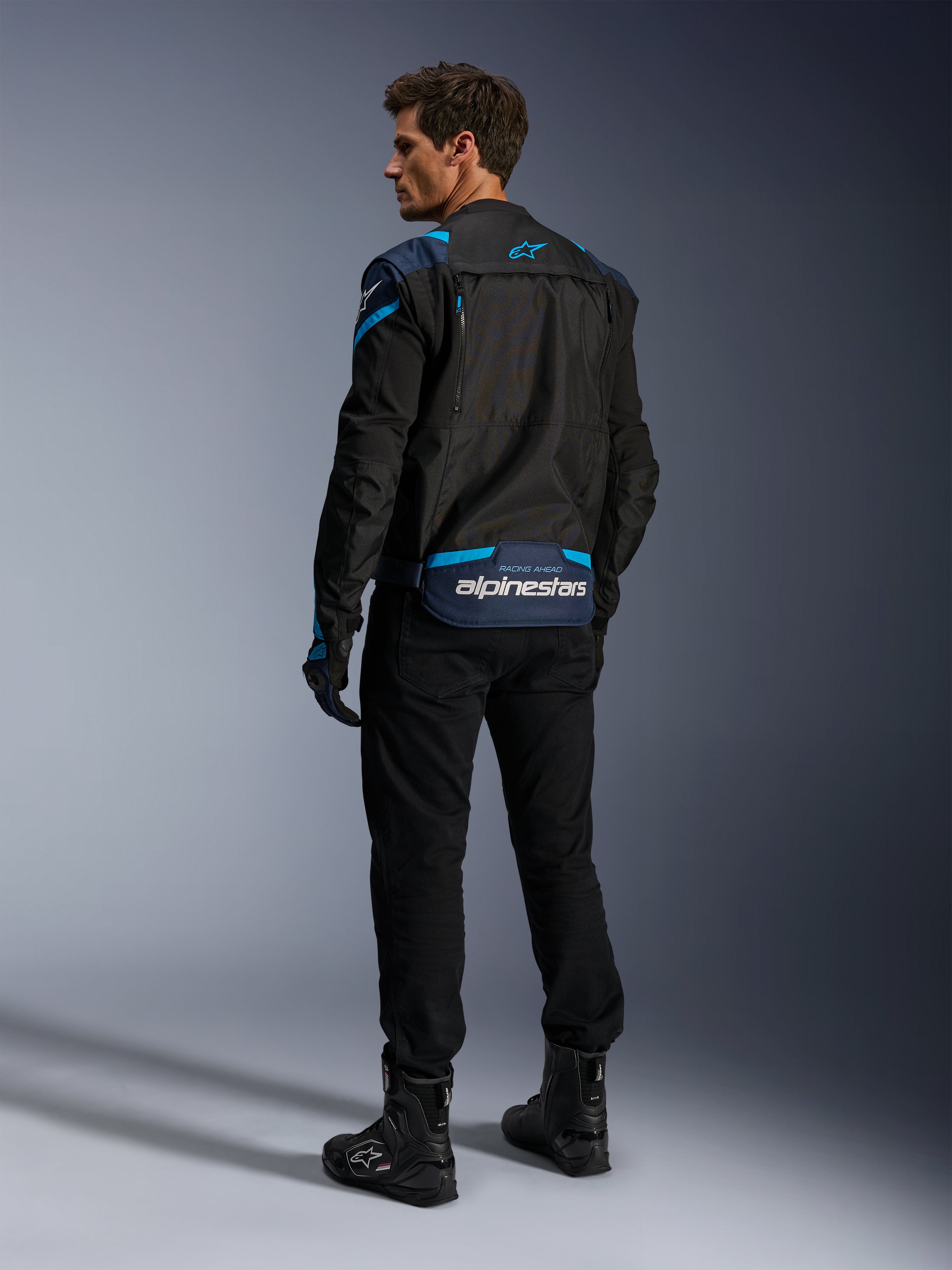Veste Alpinestars T-Stunt Air, veste de moto, noire avec accents bleu foncé et bleu vif, vue arrière montrant les panneaux en mesh modulaires et le logo sur le bas du dos, portée par un mannequin avec pantalon et bottes de moto noirs