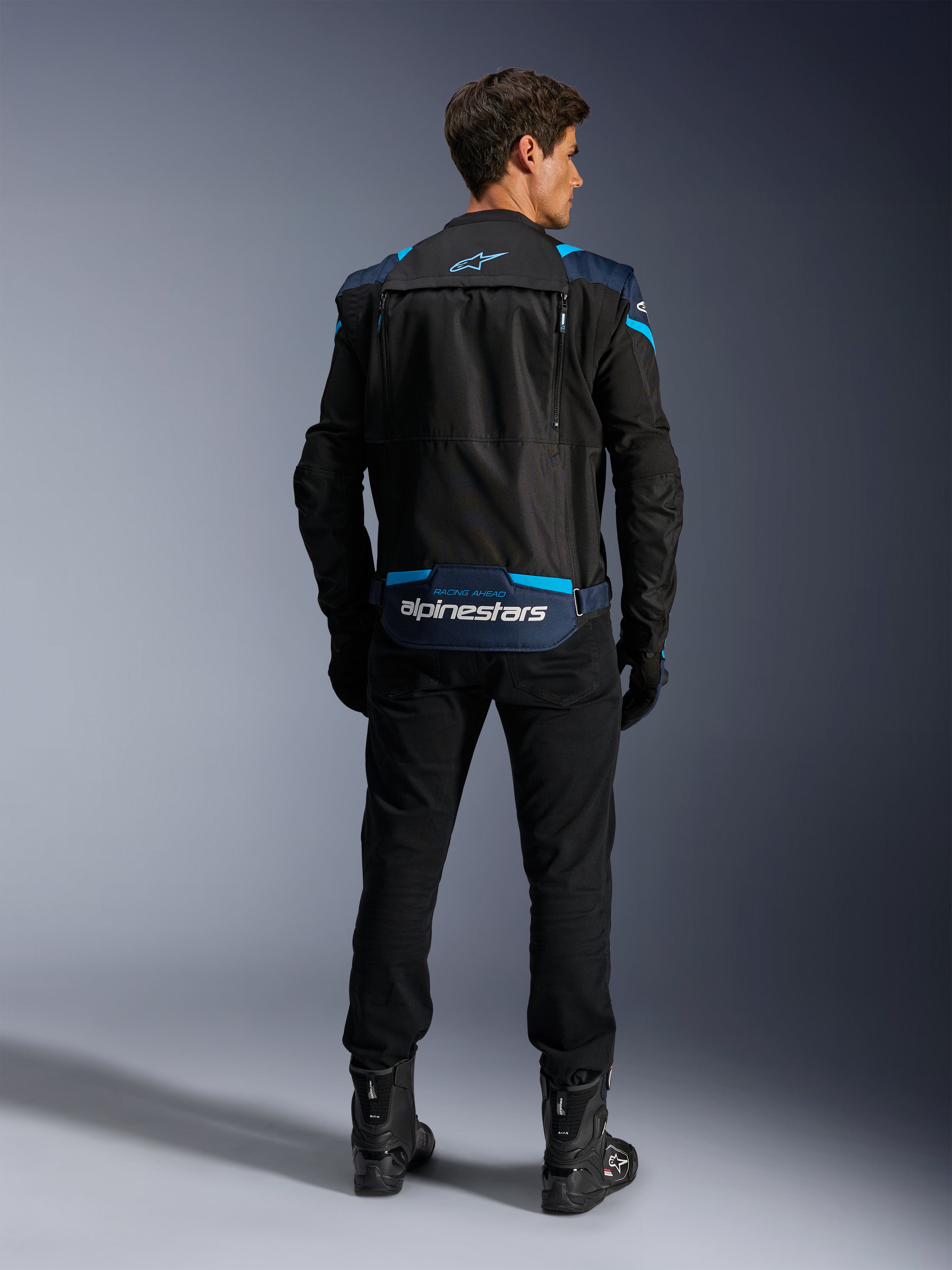 Veste Alpinestars T-Stunt Air, veste de moto textile modulaire, noir, bleu foncé et bleu vif, vue de dos montrant le compartiment de rangement des manches au bas du dos avec logo Alpinestars blanc, portée avec un pantalon et des bottes noirs