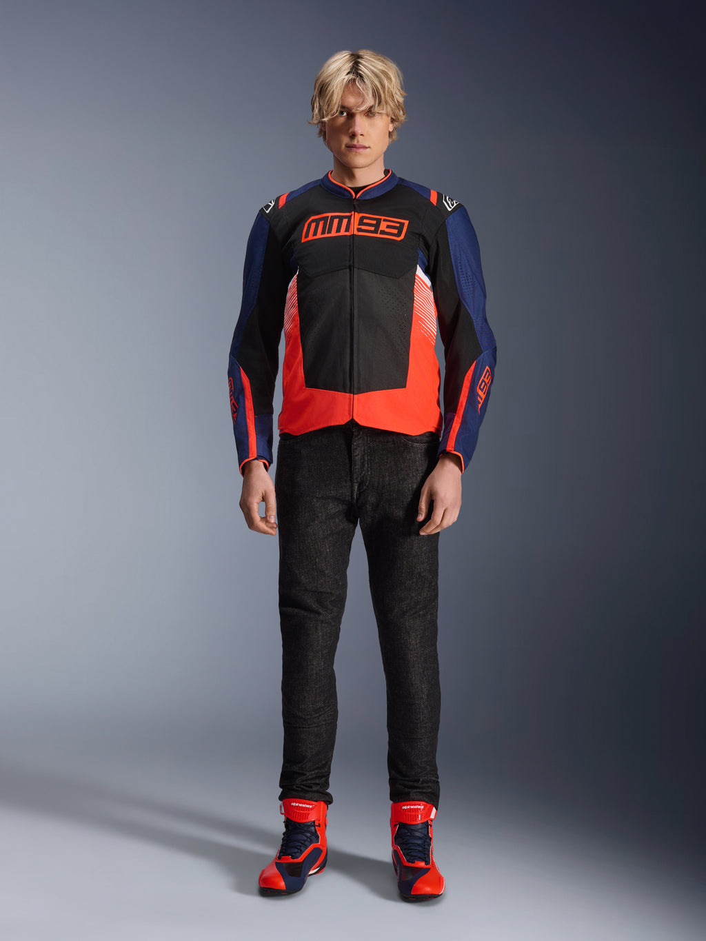 Veste Alpinestars MM93 T-SPS Air V2, bleu marine et noir avec accents rouge fluo, arborant le logo MM93 emblématique sur la poitrine, portée avec un pantalon de moto noir et des bottes assorties bleues et rouges, tenue de moto urbaine
