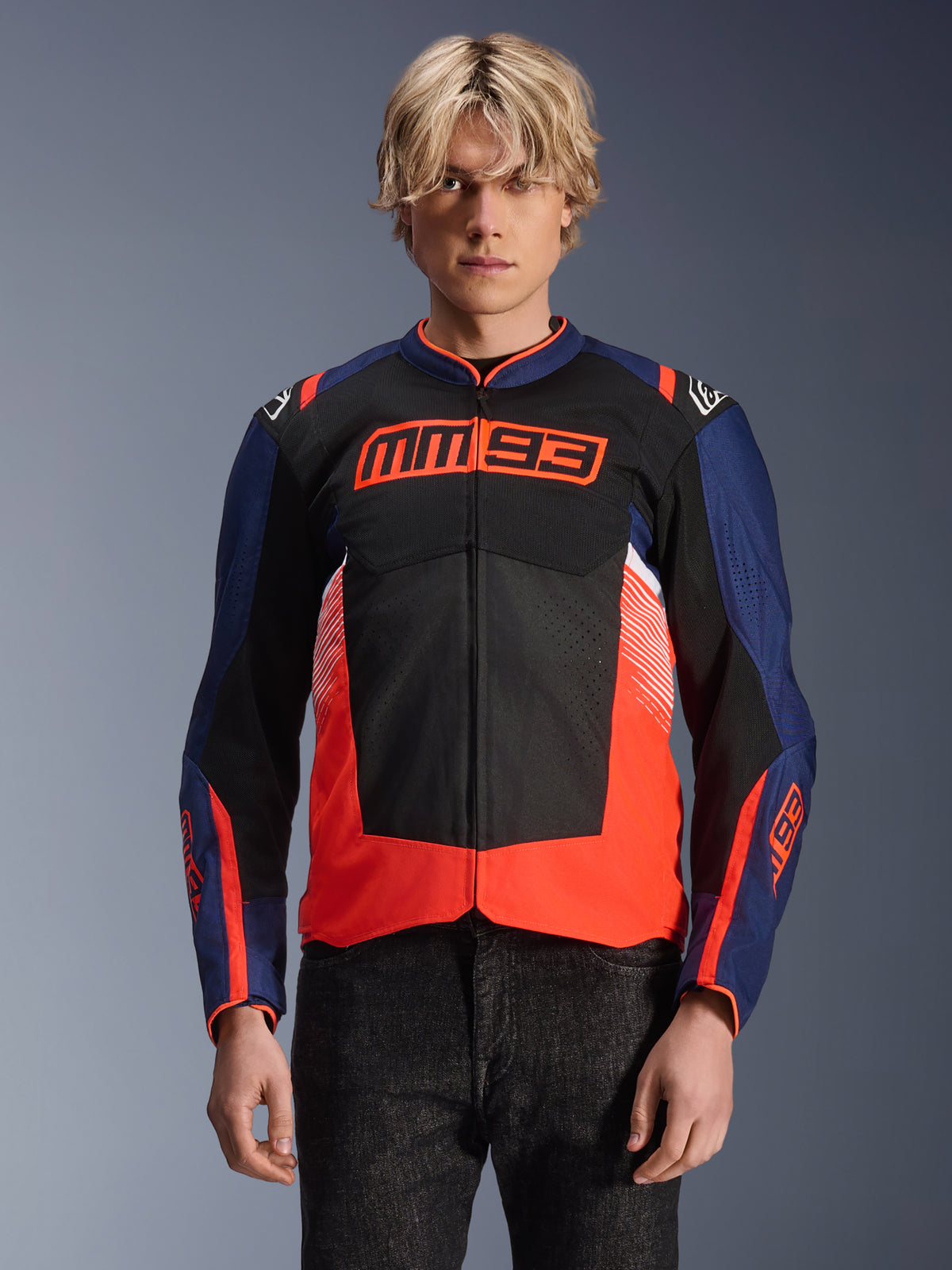 Veste Alpinestars MM93 T-SPS Air V2, veste de moto en textile, bleu marine, noir et rouge fluorescent, portée par une personne, avec un logo MM93 imposant sur la poitrine, des panneaux en mesh pour la ventilation et un design moderne à blocs de couleurs
