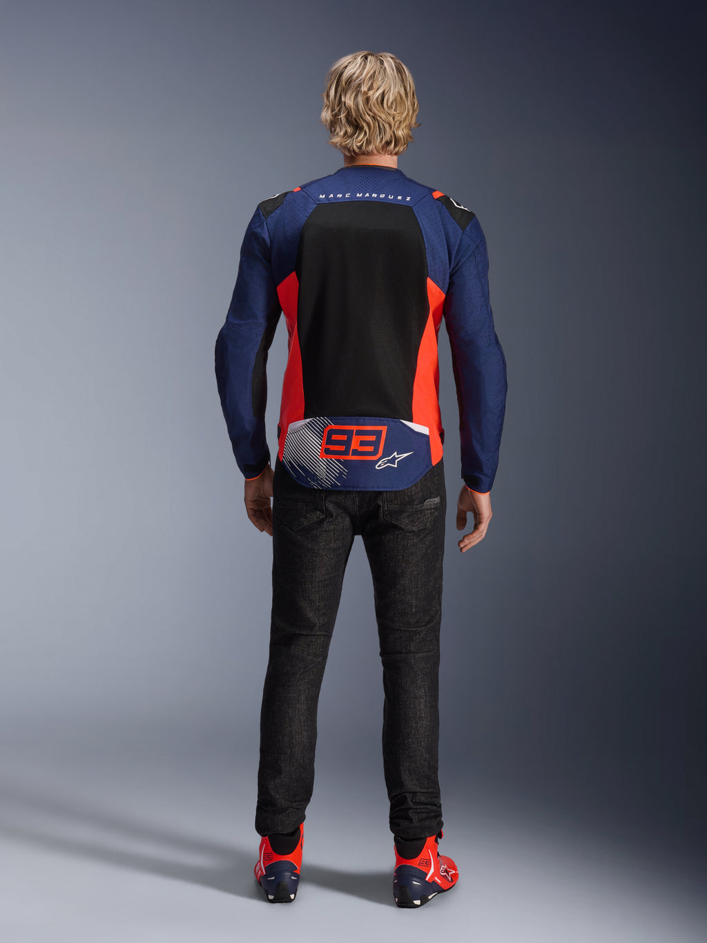 Vue arrière d'un mannequin portant la veste MM93 T-SPS Air V2 en bleu marine avec des accents rouge fluo et noirs, dotée d'un grand panneau arrière en mesh et du marquage '93' Marc Marquez, associée à un pantalon en denim foncé et des bottes de moto assorties.