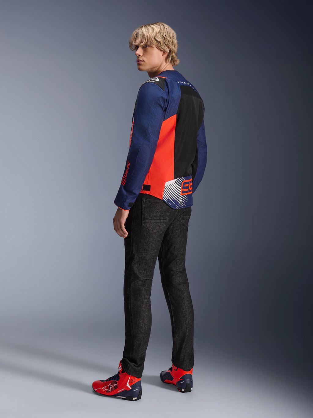 Veste Alpinestars MM93 T-SPS Air V2, bleu marine et rouge fluo, portée par un mannequin de dos avec un jean de moto noir et des chaussures assorties rouges et bleues, dotée de panneaux en mesh et du marquage Marc Márquez 93.