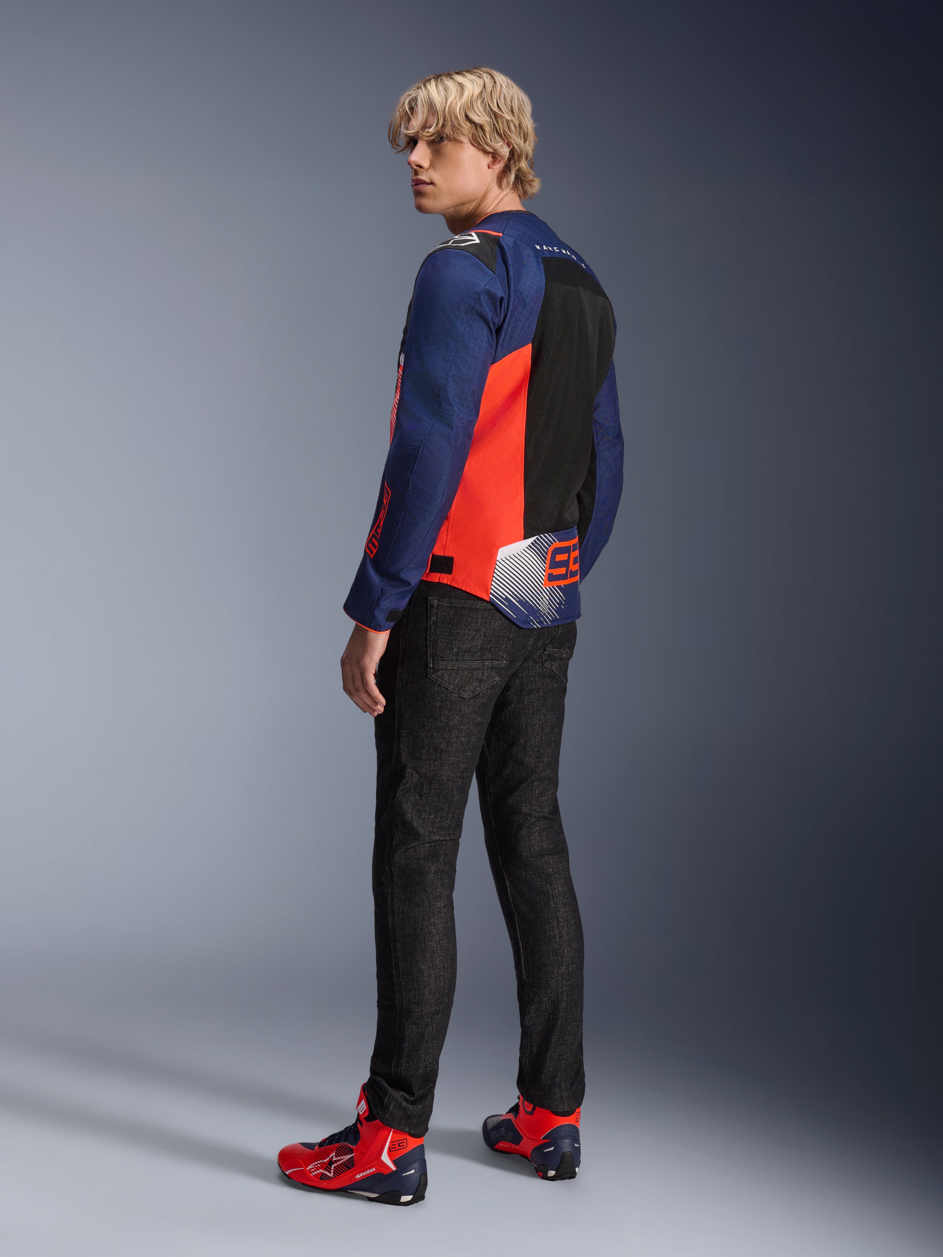 Veste Alpinestars MM93 T-SPS Air V2, bleu marine et rouge fluo, portée par un mannequin de dos avec un jean de moto noir et des chaussures assorties rouges et bleues, dotée de panneaux en mesh et du marquage Marc Márquez 93.