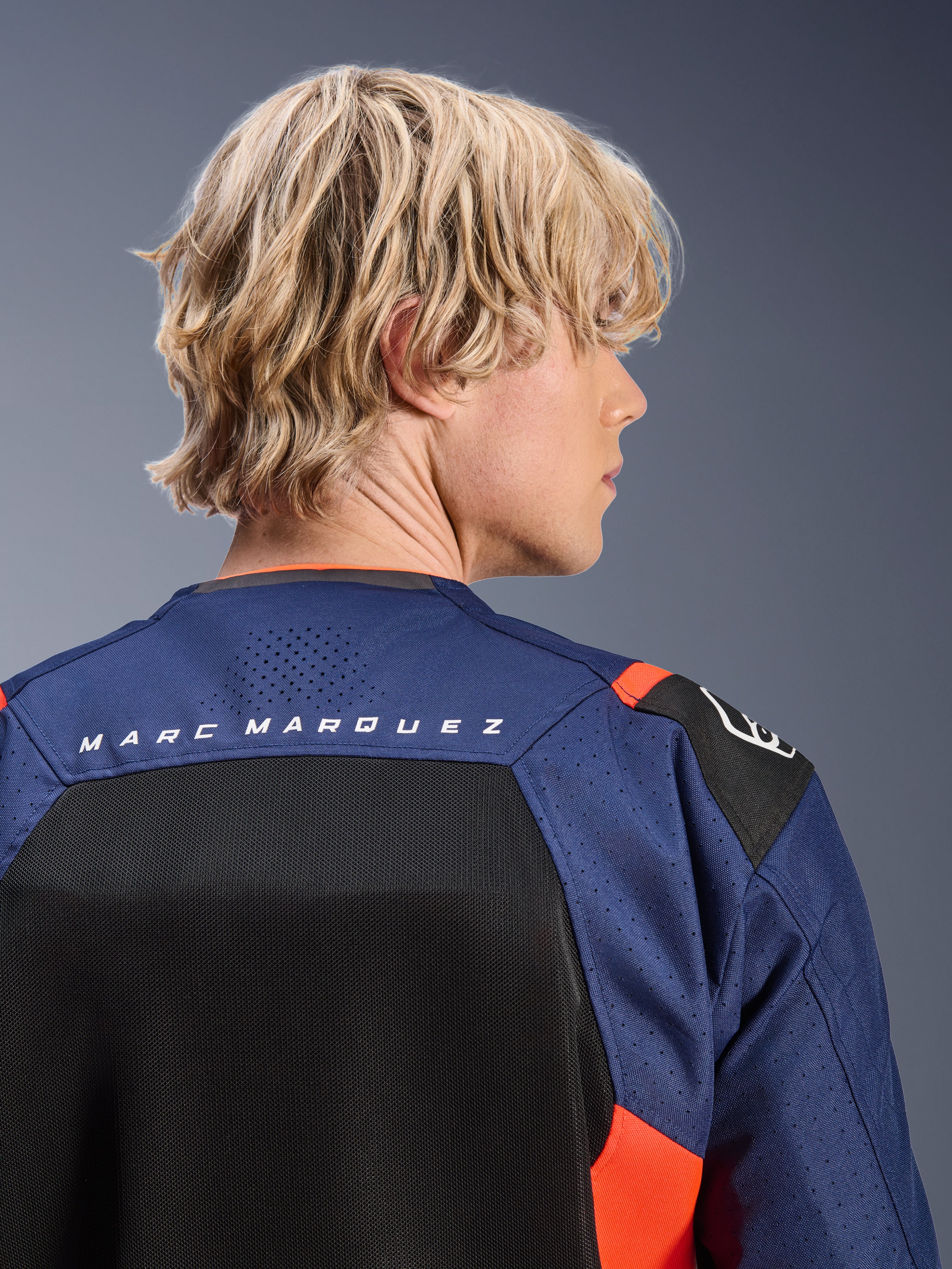 Veste Alpinestars MM93 T-SPS Air V2, bleu marine et noir avec des accents rouge fluo, gros plan sur le haut du dos arborant le logo 'MARC MARQUEZ', panneaux en mesh respirant et construction en textile perforé, portée par un mannequin blond.