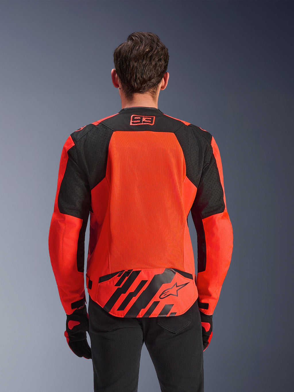 Veste Alpinestars MM93 T-SPS Air V2, vue de dos, rouge fluo et noir, avec le logo de course 93 sur le col, un grand panneau en maille dans le dos pour la ventilation, un rabat au bas du dos avec le logo étoile Alpinestars et des rayures géométriques, portée avec un pantalon noir et des gants de moto