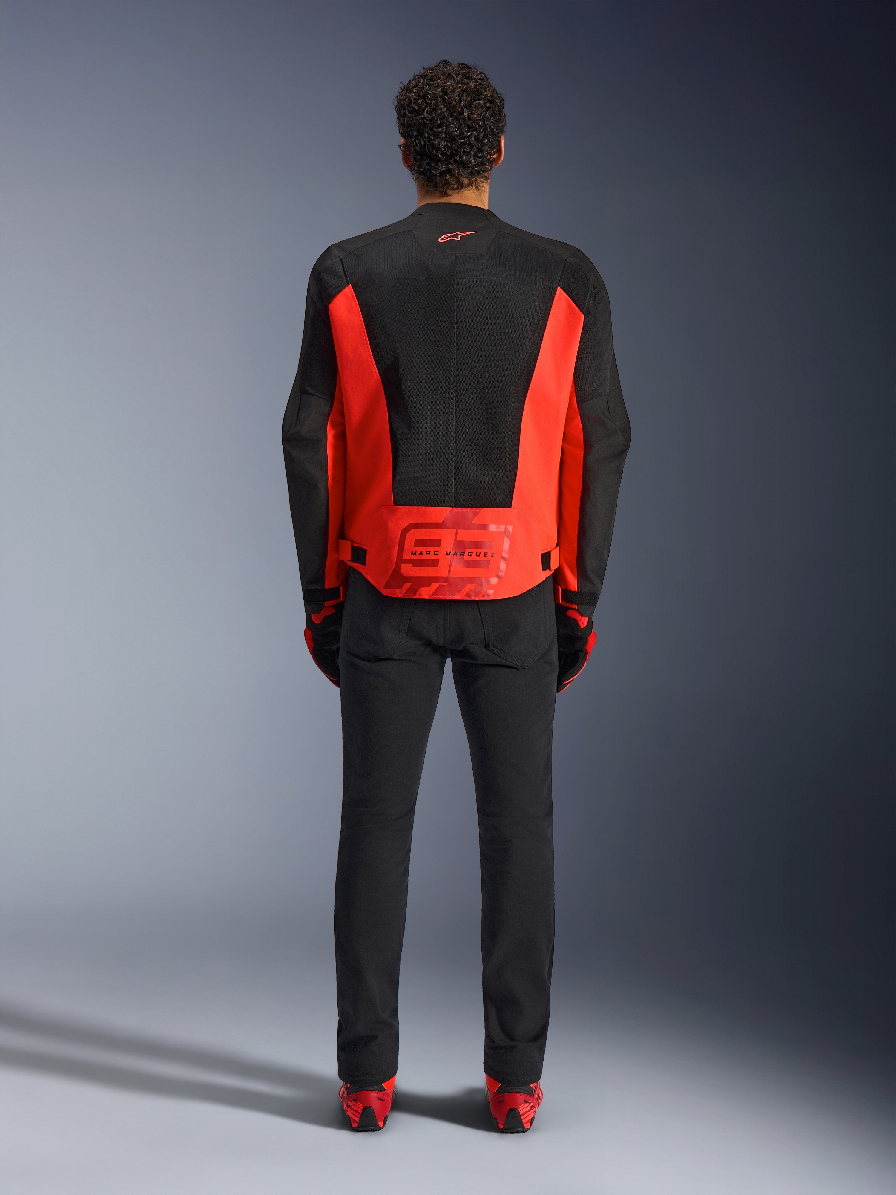 Alpinestars MM93 C-1 Air Jacket, veste de moto, rouge fluo et noir, vue arrière avec le graphisme Marc Marquez 93 sur le bas du dos, panneaux en maille technique pour la ventilation, associée à un pantalon noir et un équipement aux accents rouges