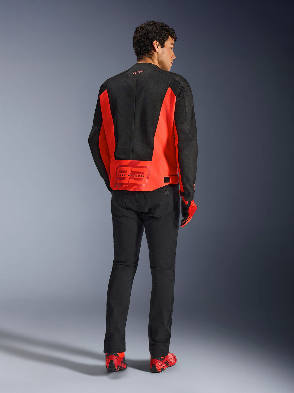 Vue arrière d'un mannequin portant la veste Alpinestars MM93 C-1 Air en rouge fluo et noir, avec le logo Marc Marquez 93 sur le bas du dos, associée à un pantalon noir et des chaussures de moto rouges, style urbain pour motards