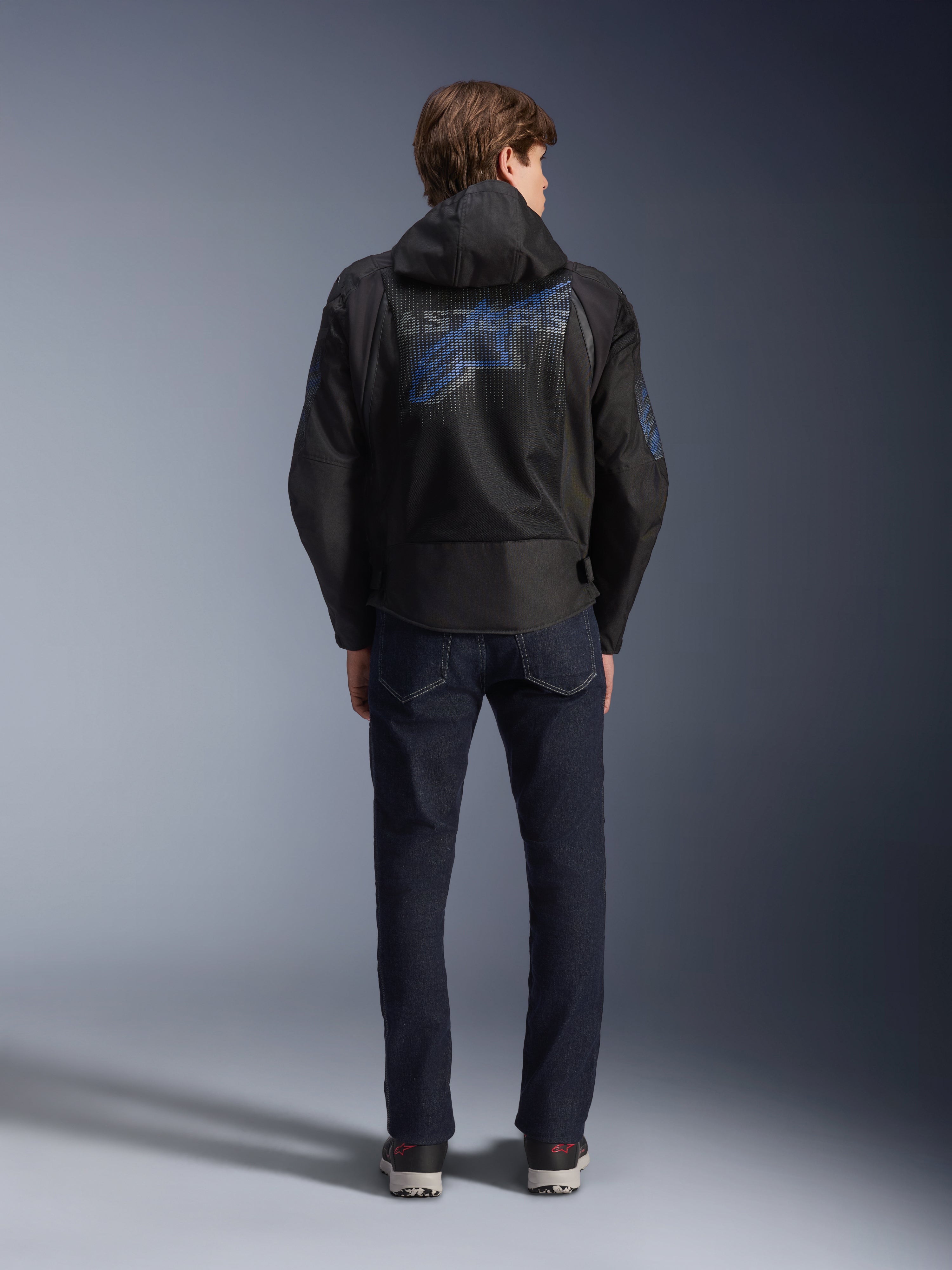 Alpinestars Zaca Air Venom WP Jacket, Black and Electric Blue, rear view featuring a large blue Alpinestars logo on the back mesh panel, detachable hood, worn with dark denim jeans and sneakers, sporty all-weather motorcycle gear.