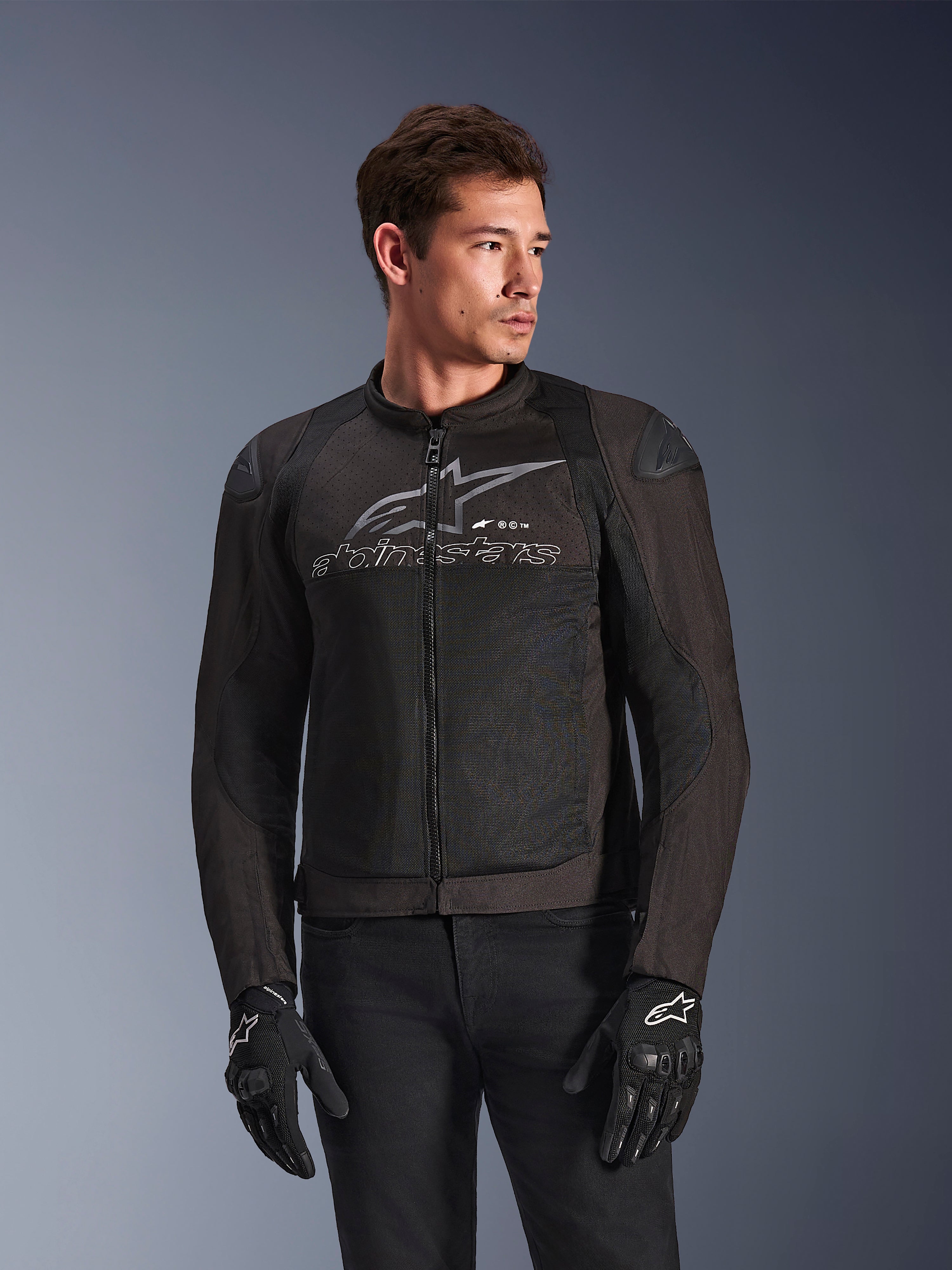 Veste Alpinestars SMX Air en Noir Anthracite, veste de moto en textile avec panneaux en maille à flux élevé, dotée d'un grand logo et texte Alpinestars gris sur la poitrine, sliders d'épaule DFS Lite, portée par un mannequin avec des gants de moto noirs, style de conduite sportive.
