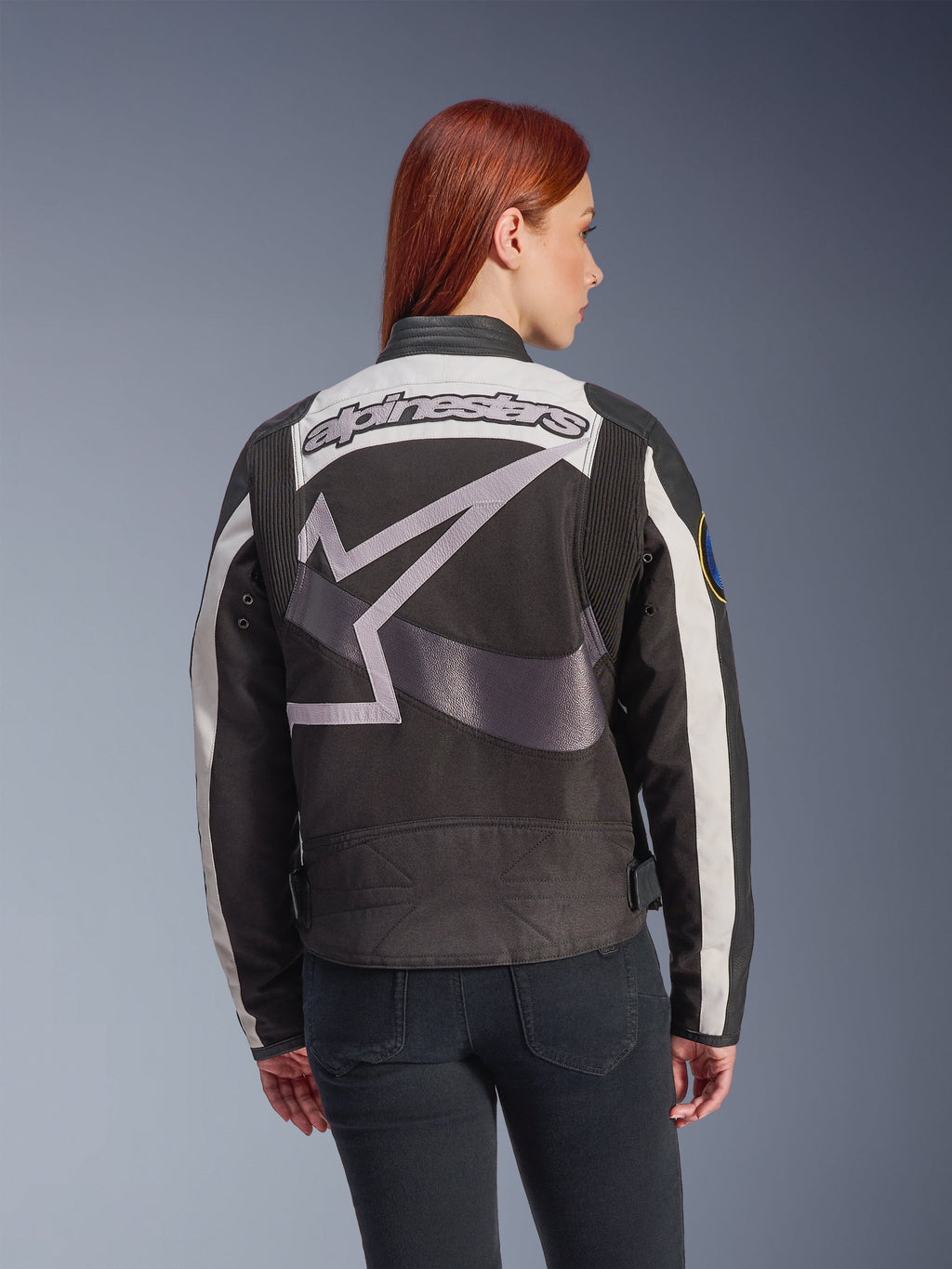 Veste Alpinestars Idles pour femme, noir et blanc cassé, vue de dos avec un grand logo étoile gris et le lettrage Alpinestars, portée par un mannequin aux cheveux roux, veste de moto urbaine d'inspiration vintage avec panneaux en accordéon.