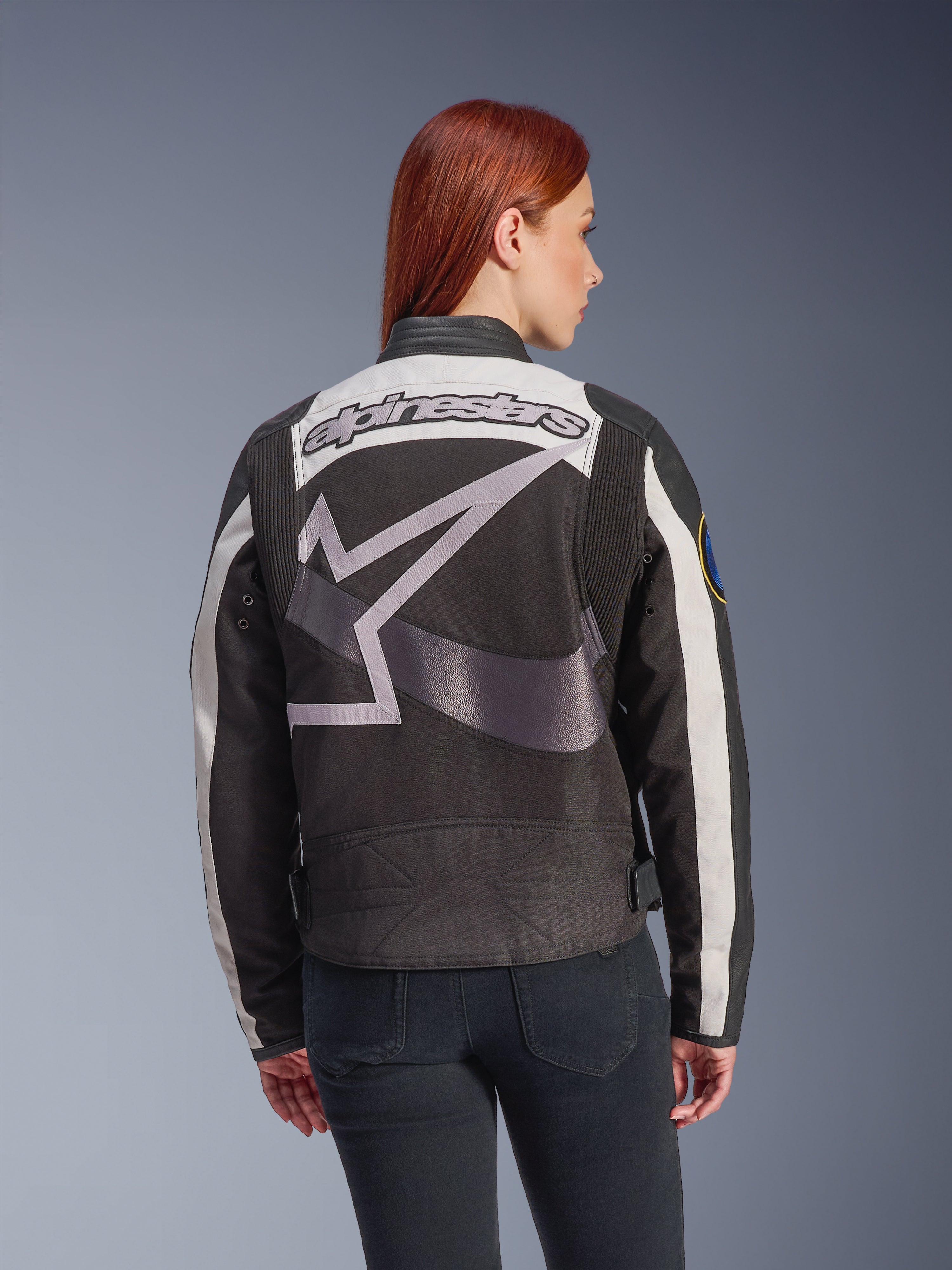 Veste Alpinestars Idles pour femme, noir et blanc cassé, vue de dos avec un grand logo étoile gris et le lettrage Alpinestars, portée par un mannequin aux cheveux roux, veste de moto urbaine d'inspiration vintage avec panneaux en accordéon.