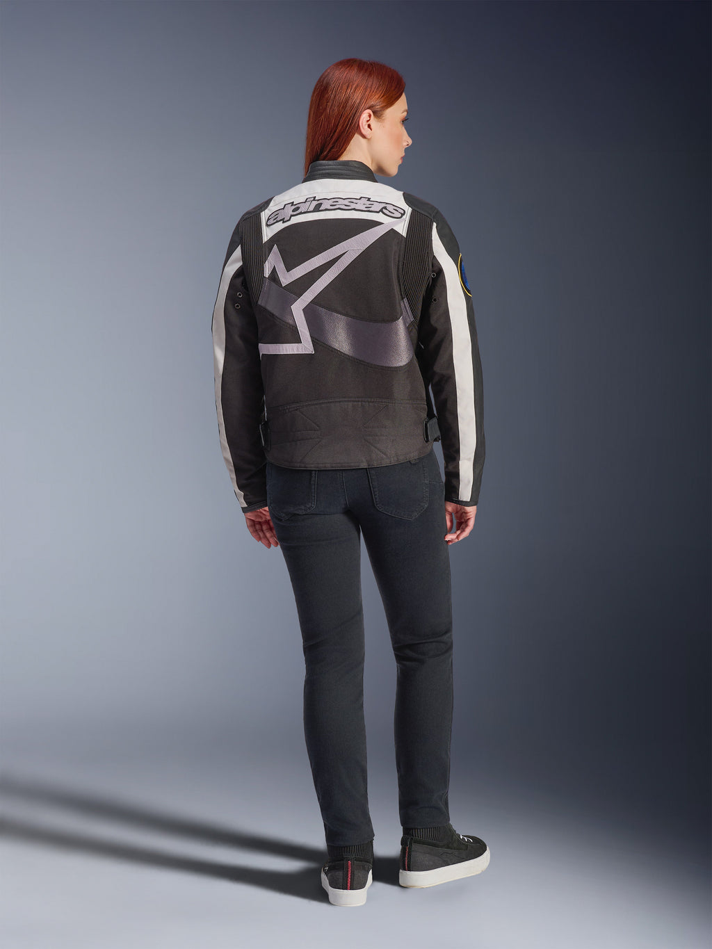 Veste Alpinestars Idles pour femme, veste de moto urbaine, noir et blanc cassé, vue de dos présentant un large logo et un mot-symbole Alpinestars gris, portée avec un jean noir slim et des baskets, design d'inspiration vintage pour la conduite urbaine