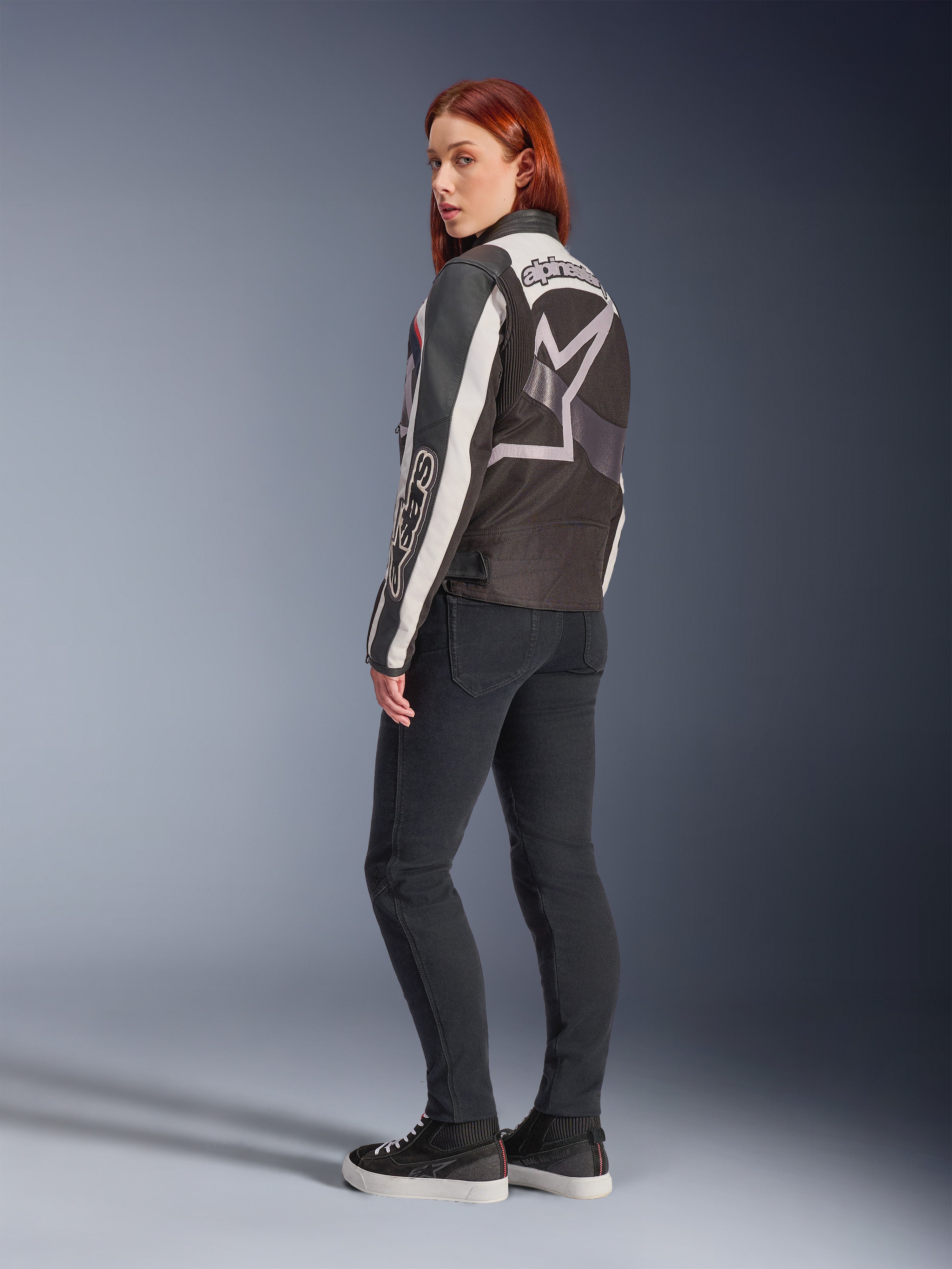 Femme portant la veste Alpinestars Idles, vue de dos, coloris blanc cassé, rouge flamme et noir, avec un grand logo étoilé dans le dos et l'inscription Alpinestars sur la manche, accompagnée d'un pantalon slim noir et de baskets, vêtement de moto urbain pour femme