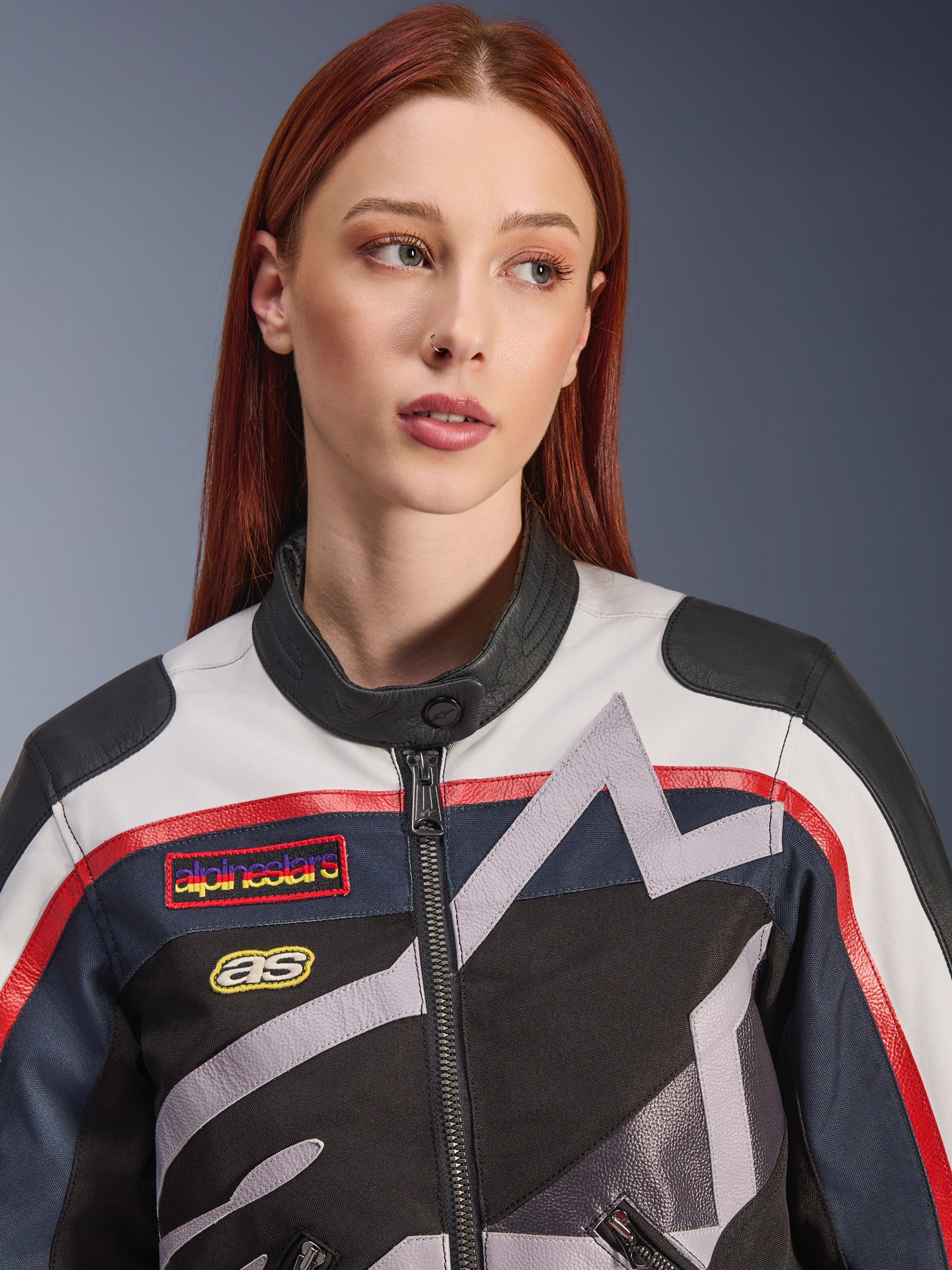 Veste femme Alpinestars Idles, gros plan d'un mannequin aux cheveux roux, coloris blanc cassé, rouge et noir, design de course d'inspiration vintage avec écussons et graphismes d'étoiles géométriques grises, équipement de moto urbain