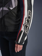 Veste pour femme Alpinestars Idles, veste de moto urbaine, blanc cassé, rouge flamme et noir, gros plan sur la manche avec un grand logo Alpinestars brodé, bandes de course rouges et blanches, construction en textile et cuir noirs, et détails de fermeture éclair en métal.