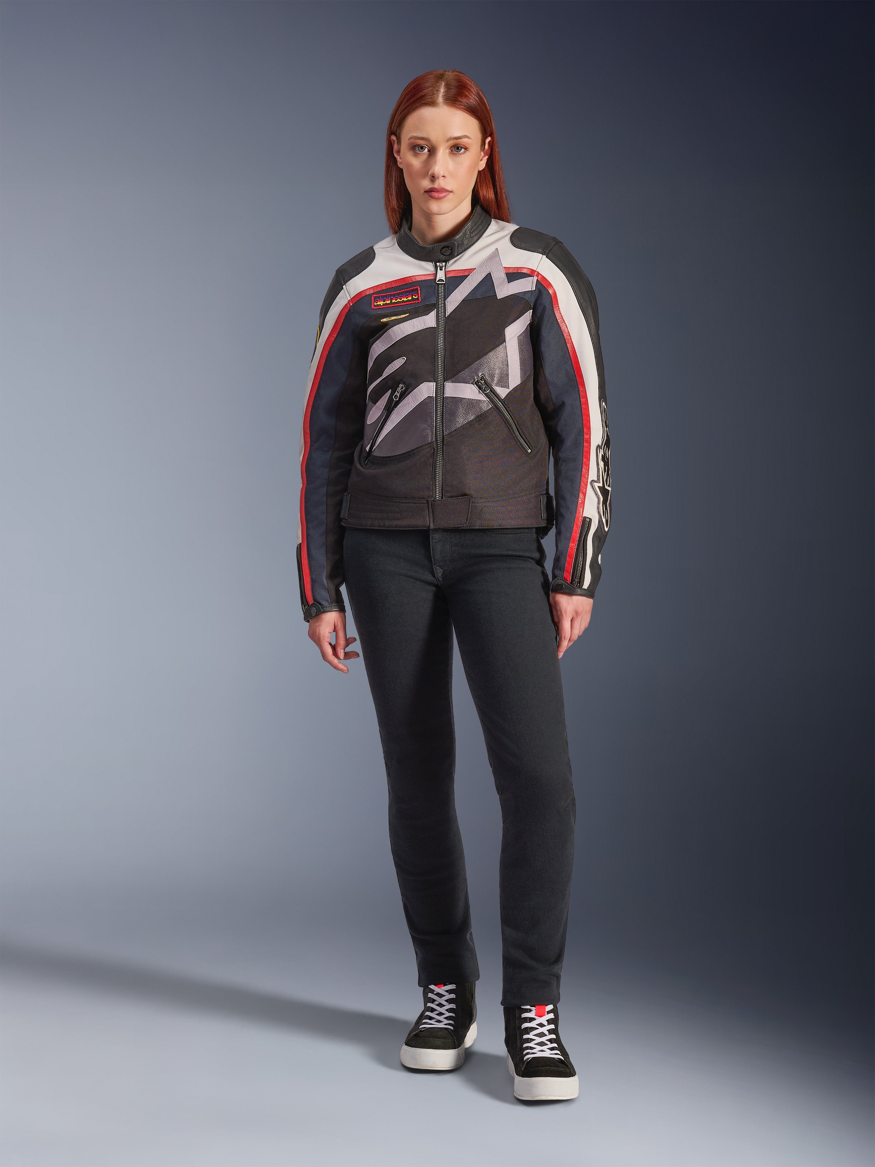 Modèle féminin portant la veste Alpinestars Idles, veste de moto pour femme, blanc cassé, rouge flamme et noir, avec un large logo étoilé enveloppant et un marquage d'inspiration vintage, associée à un pantalon de moto ajusté sombre et des baskets montantes noires, équipement de moto urbain contemporain