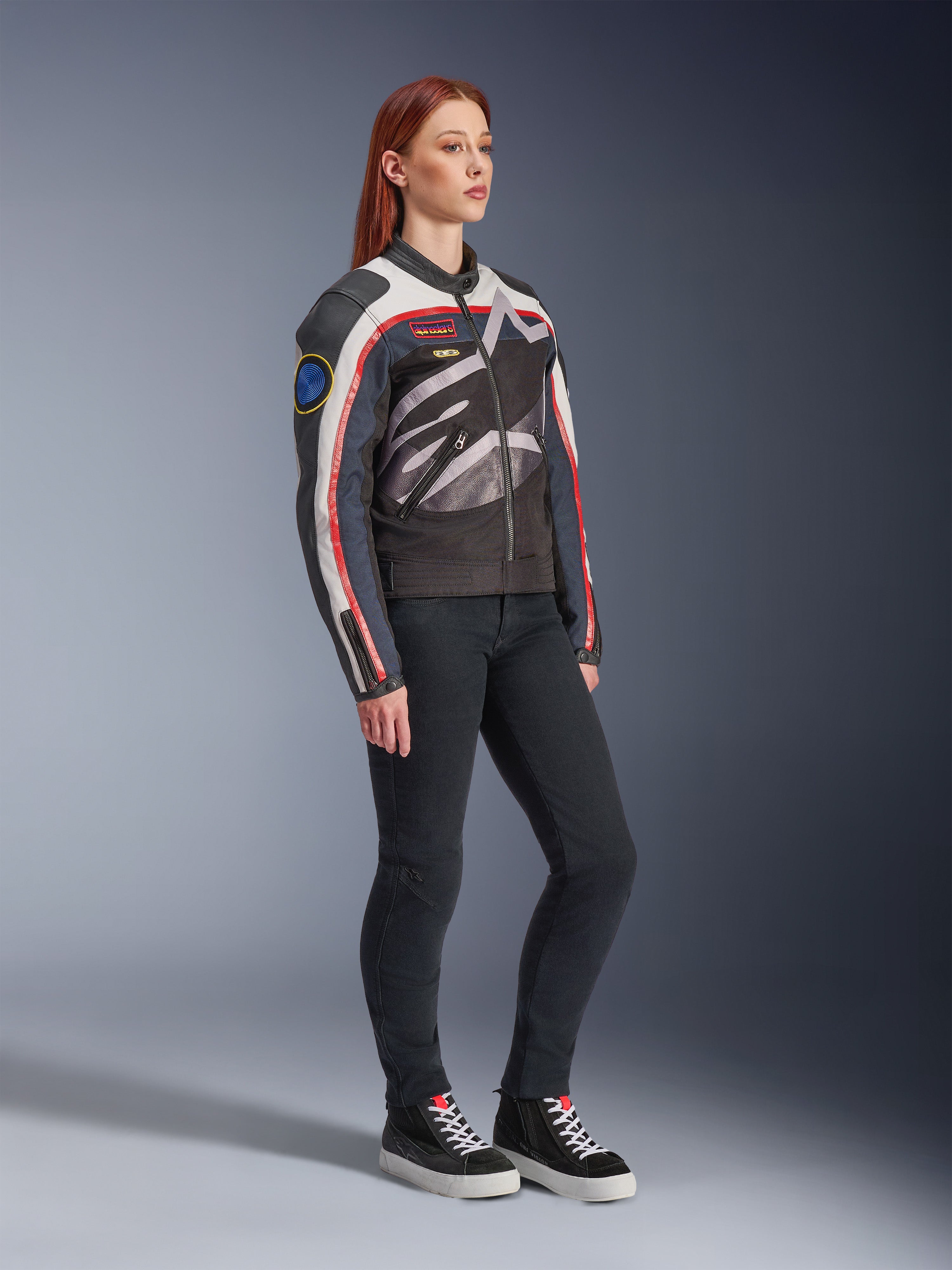Veste Alpinestars Idles pour femme, veste de moto, blanc cassé, rouge flamme et noir, arborant un grand logo Alpinestars enveloppant et des patchs d'inspiration racing vintage, portée avec un pantalon noir et des baskets montantes, équipement de conduite urbain contemporain