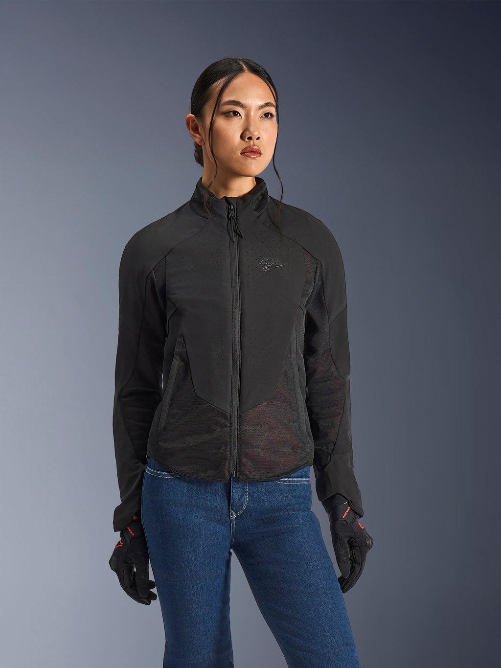 Veste Alpinestars Stella Aeroshell Airflow, veste de moto pour femme, noir anthracite, portée par un mannequin avec un jean bleu et des gants noirs, dotée d'un softshell perforé au laser et de panneaux en maille pour une ventilation optimale, design ergonomique pour la conduite sportive.