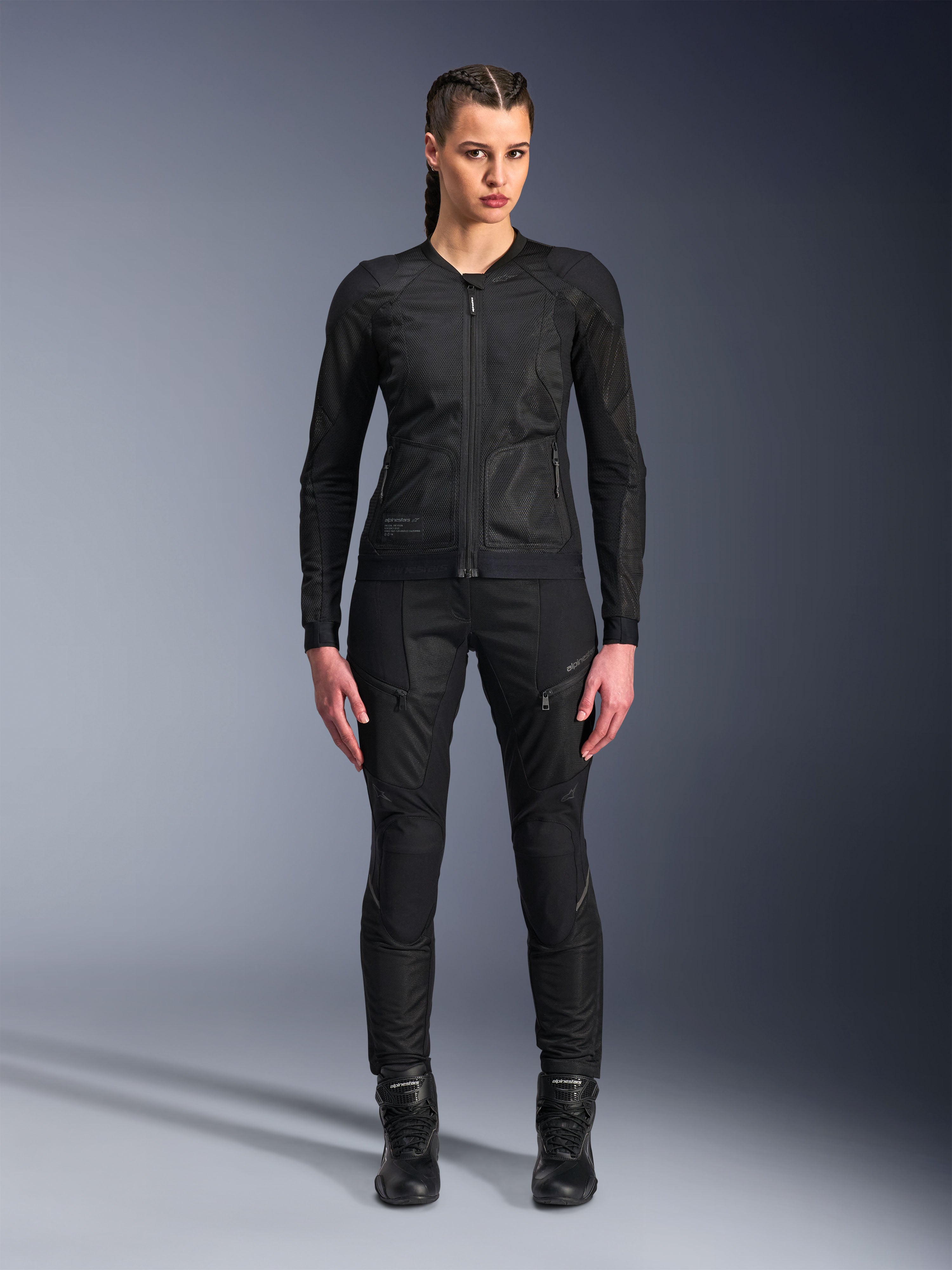 Veste Alpinestars Stella Troop-Air, veste de moto pour femme, noire, construction technique en maille 2D avec un design minimaliste, portée avec un pantalon de moto et des bottes noirs, style de moto urbain contemporain.