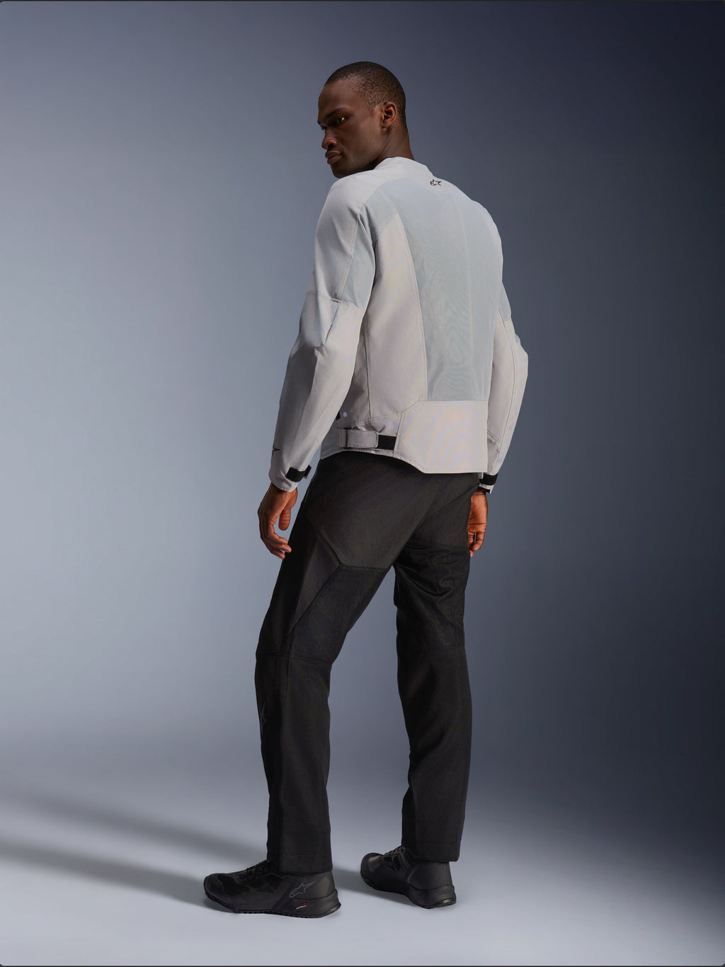 Veste Alpinestars C-1 Air, argent et gris, vue de dos sur un modèle, avec panneaux en maille technique ventilée et logos réfléchissants, portée avec un pantalon et des chaussures noirs, équipement de moto urbain contemporain.